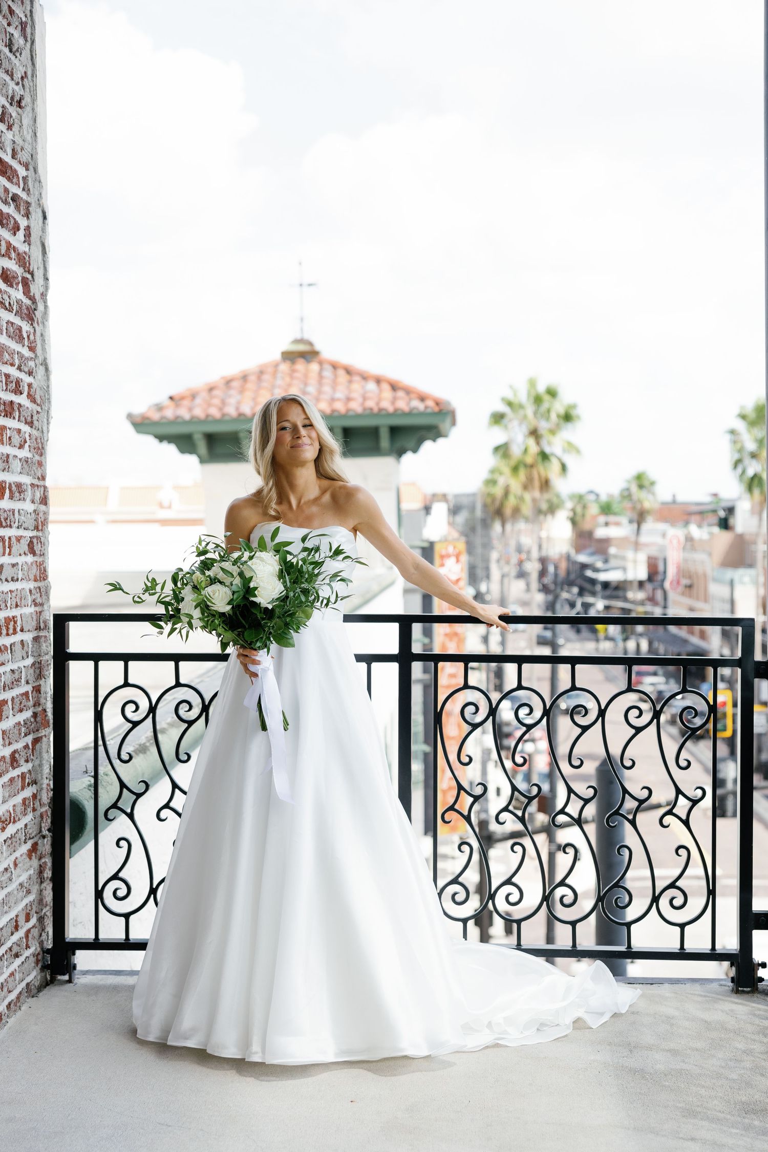 Blonde bride in strapless ballgown holds white floral bouquet on ornate iron balcony with city view.