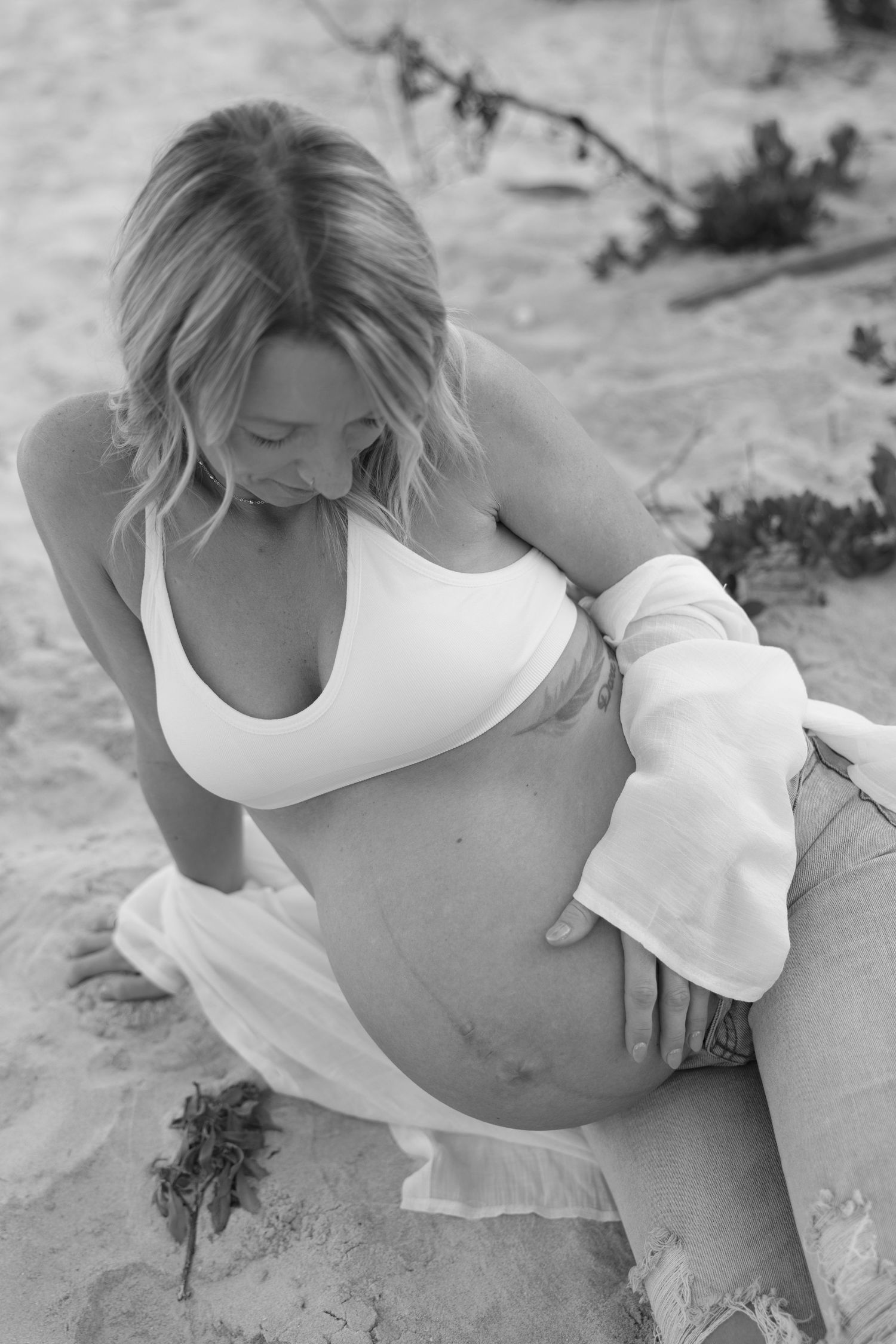 Black and white photo of pregnant woman in bikini top sitting on sand, holding her belly.