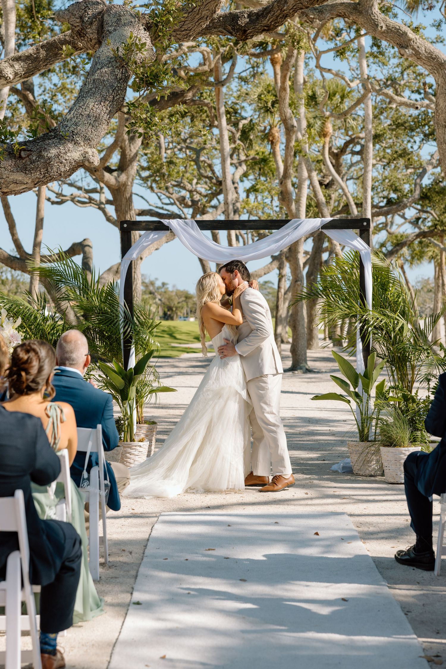 Bride and groom sharing first kiss at outdoor ceremony altar draped with white fabric under canopy of trees.