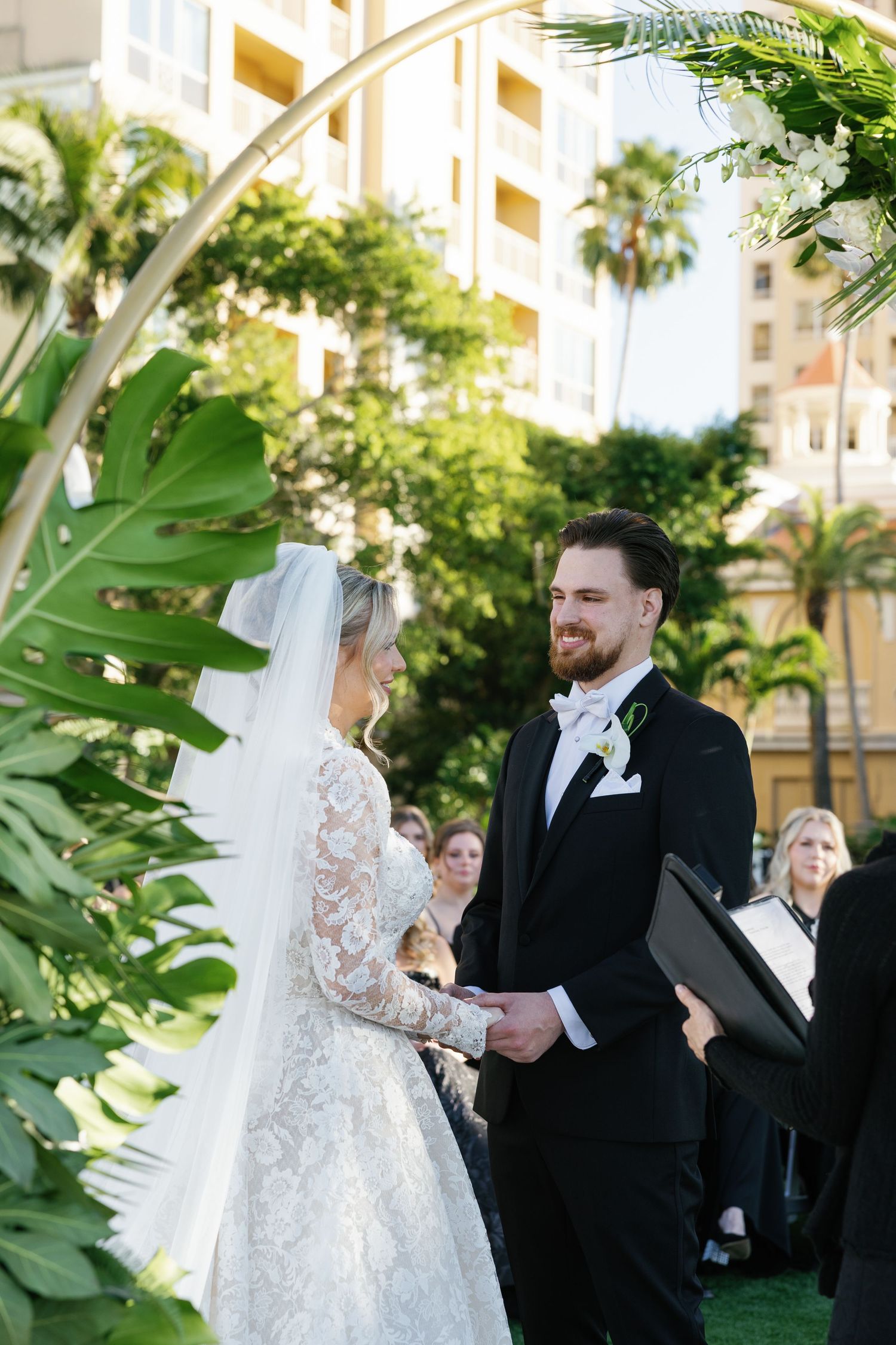 Bride and groom exchange vows outdoors at a tropical wedding ceremony, surrounded by lush greenery and palm trees.