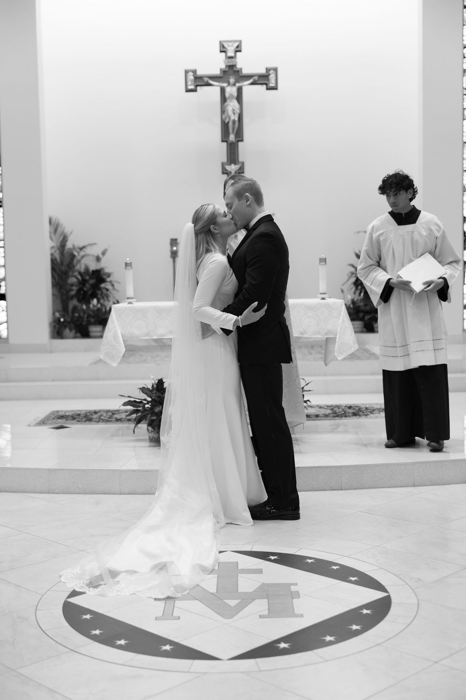 Bride and groom share first kiss at church altar during Catholic wedding ceremony in black and white photo.