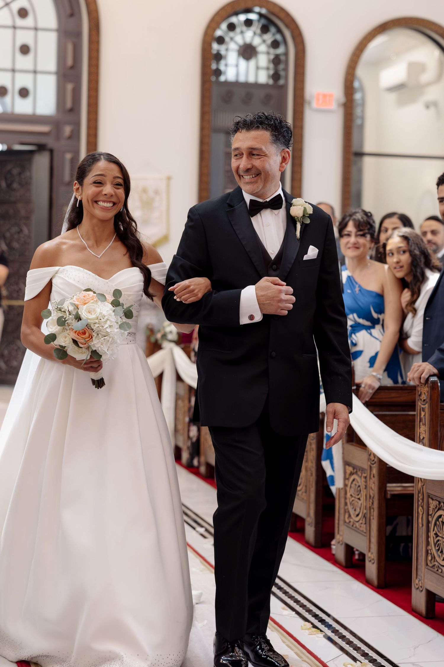 Bride in off-shoulder gown walks down red-carpeted church aisle with escort during wedding ceremony.