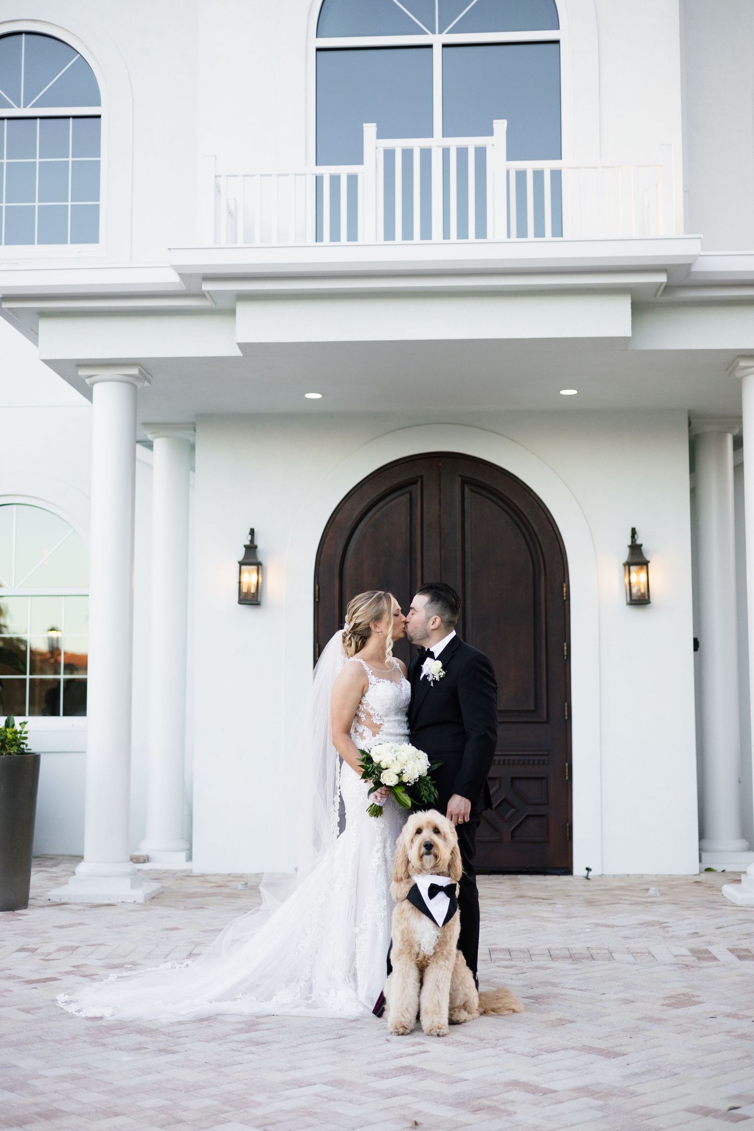 Bride and groom kiss outside elegant white venue with golden doodle dog in matching tuxedo.