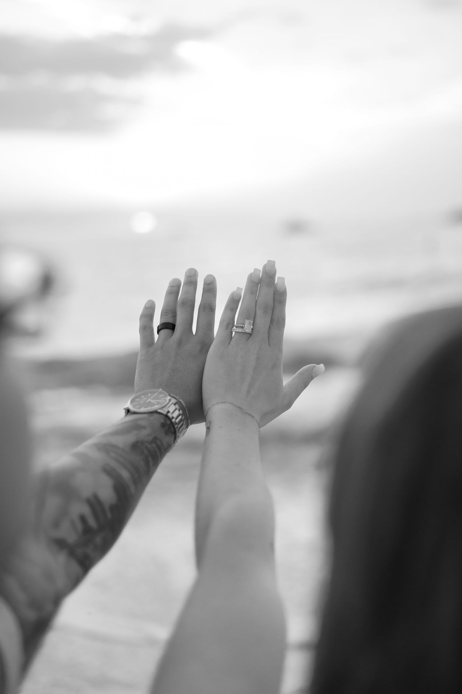 Black and white close-up of newlyweds showing wedding rings with ocean backdrop.