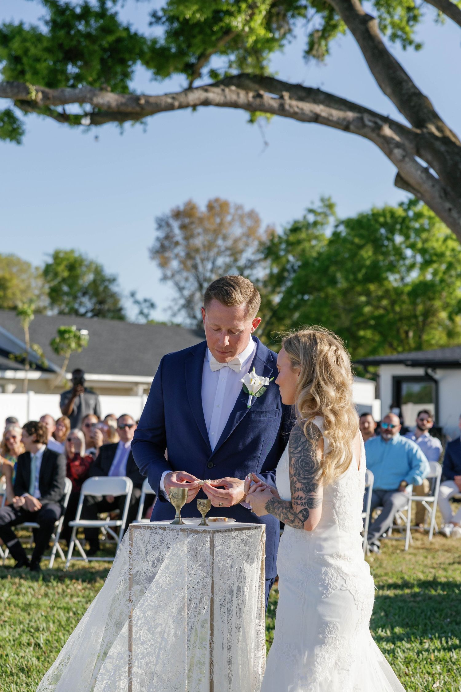 Bride and groom perform unity ceremony at outdoor wedding with tattooed bride in lace gown and groom in navy suit.