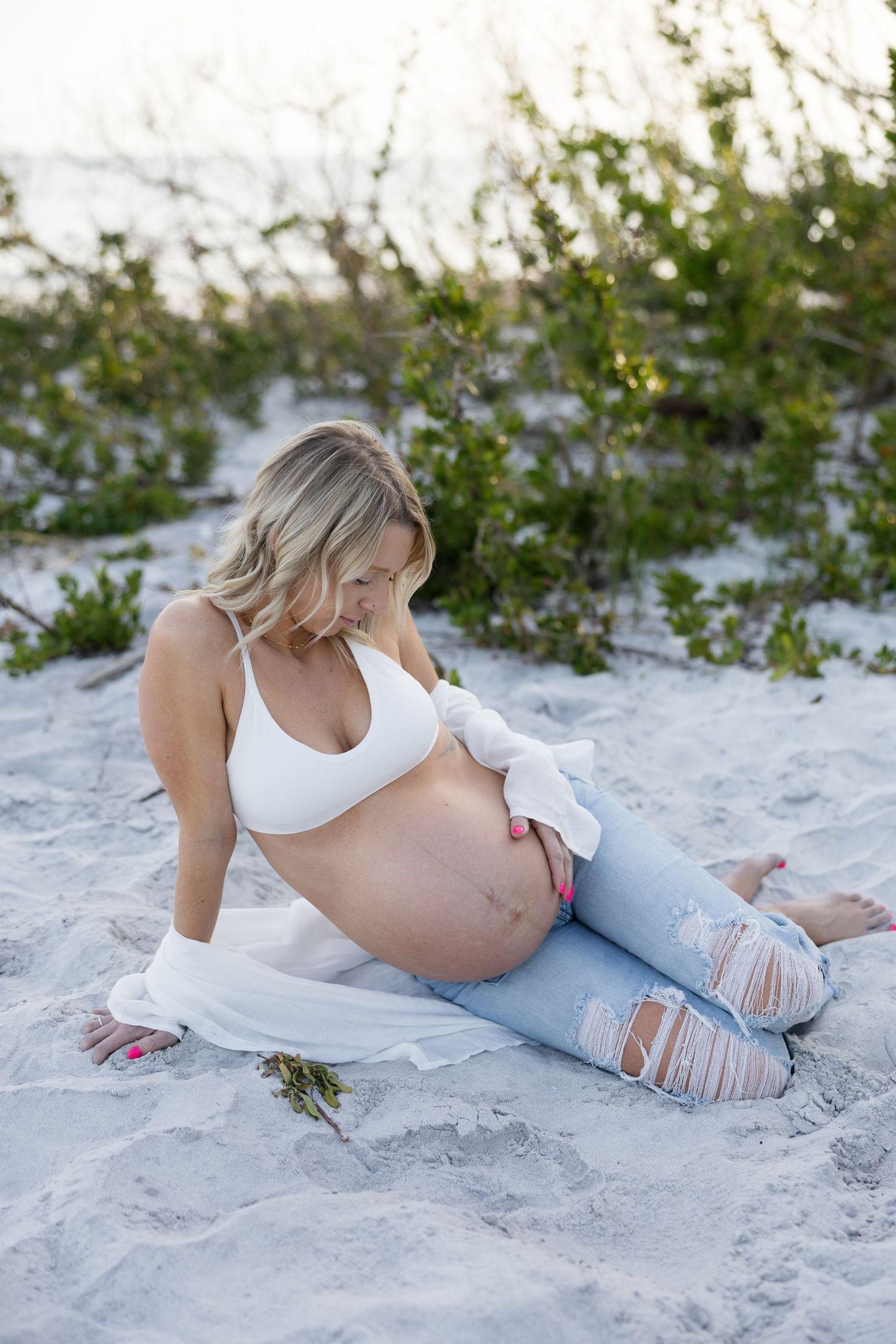 Pregnant woman in white bralette and ripped jeans sitting on sandy beach looking at belly.