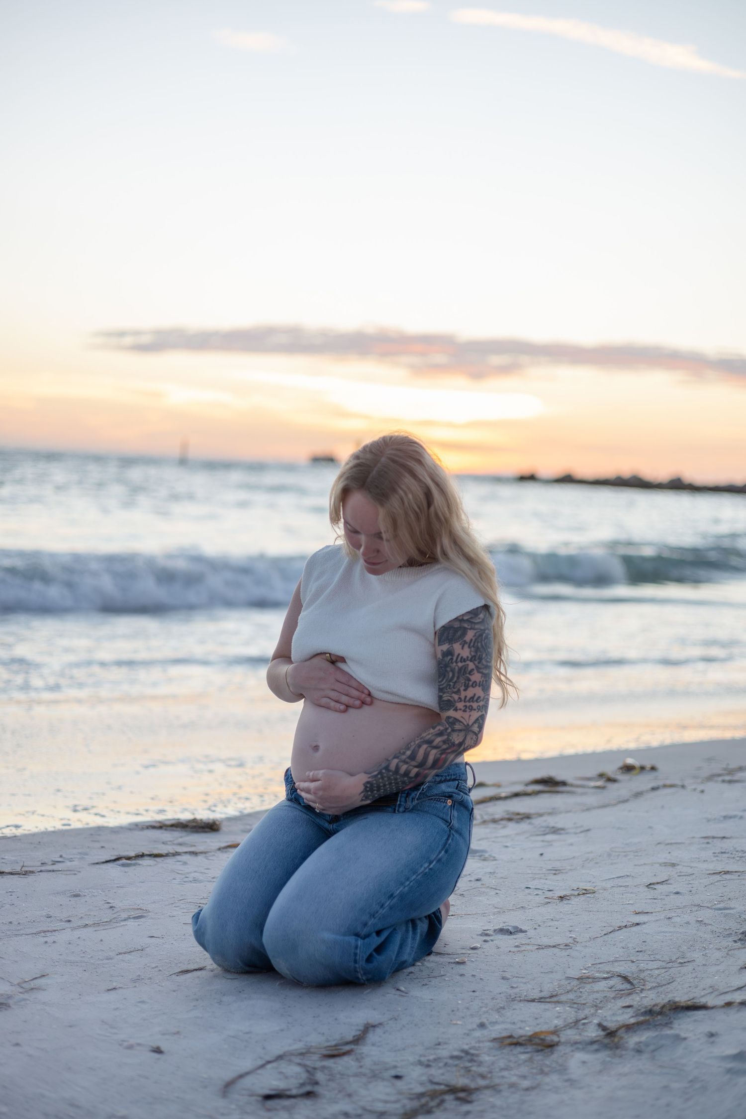 Pregnant woman kneeling on beach at sunset, cradling her bare belly with tattooed arms.
