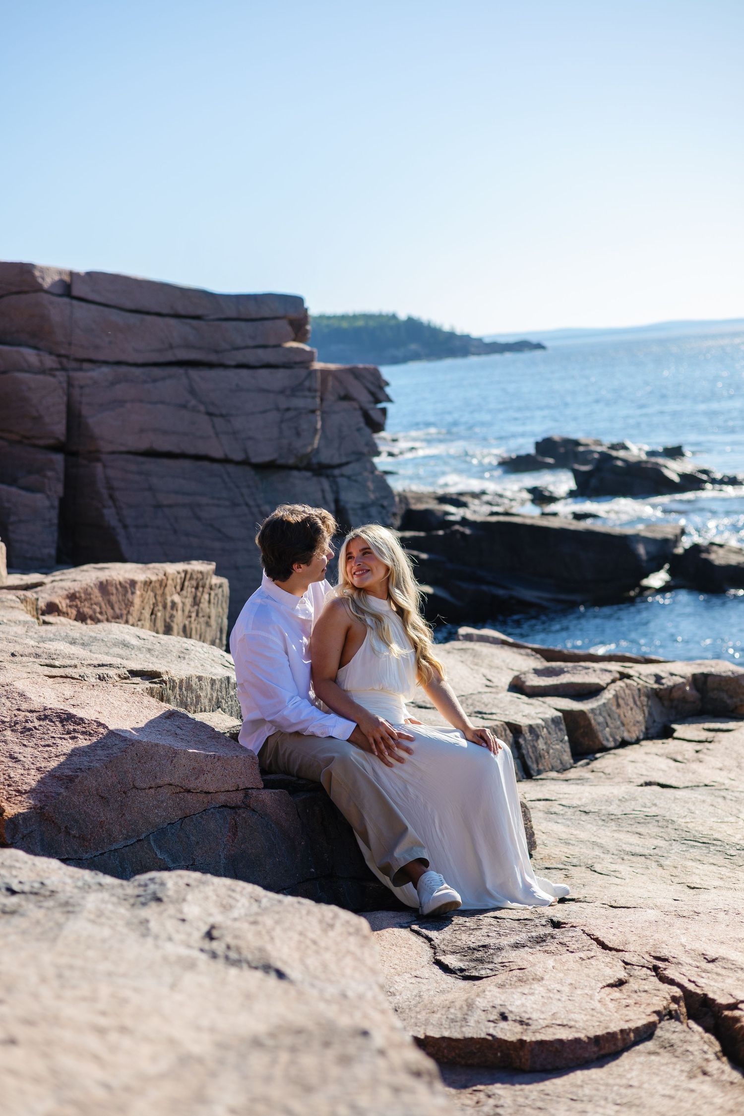 Couple sitting closely together on coastal rocks with scenic ocean and rocky shoreline backdrop.