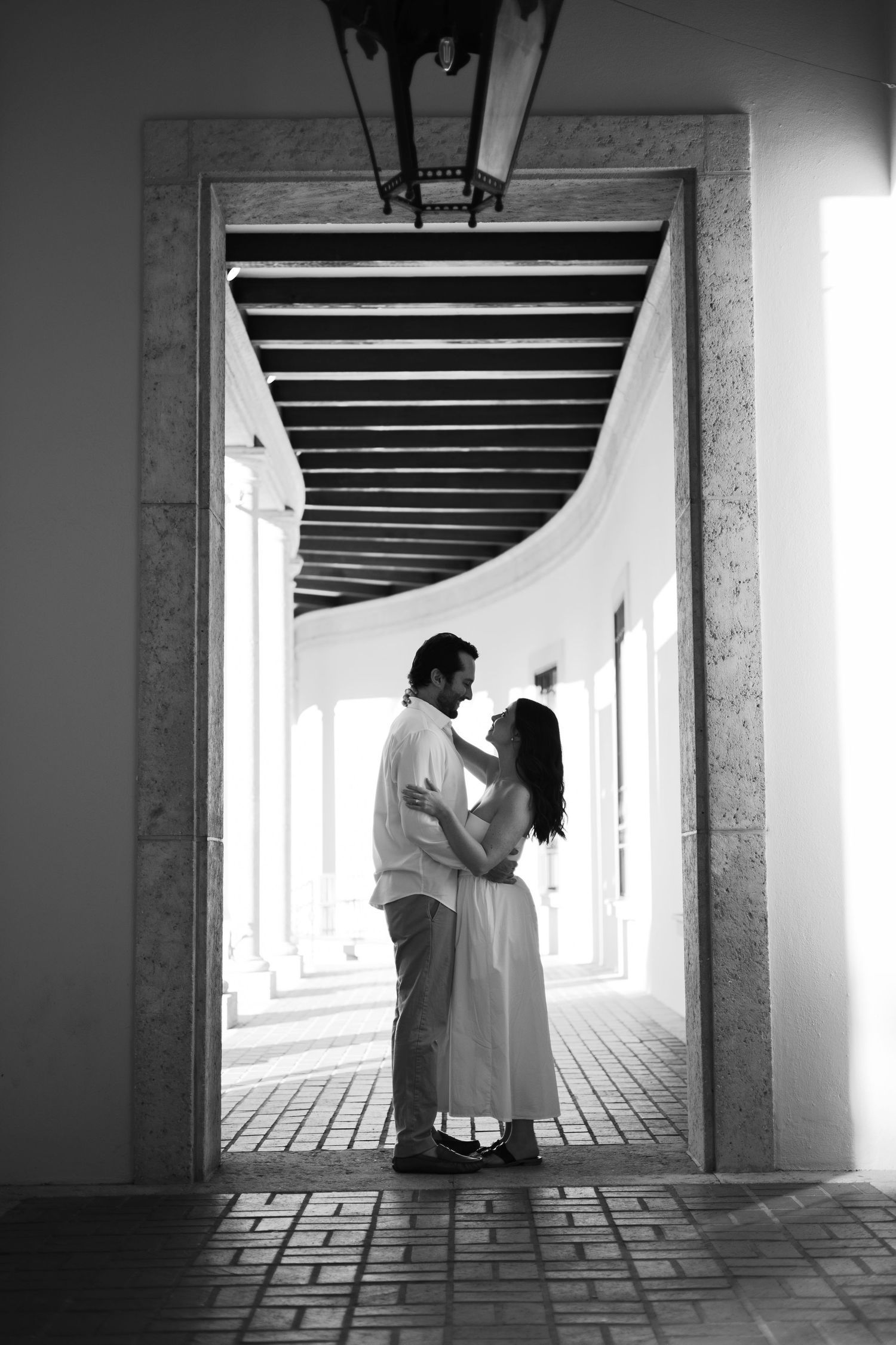 Black and white photo of romantic couple embracing in elegant arched corridor.