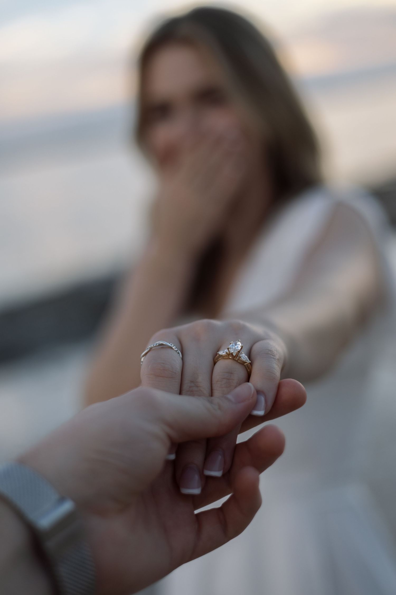 Close-up of an engagement ring on woman's hand held by man, with surprised bride in background.