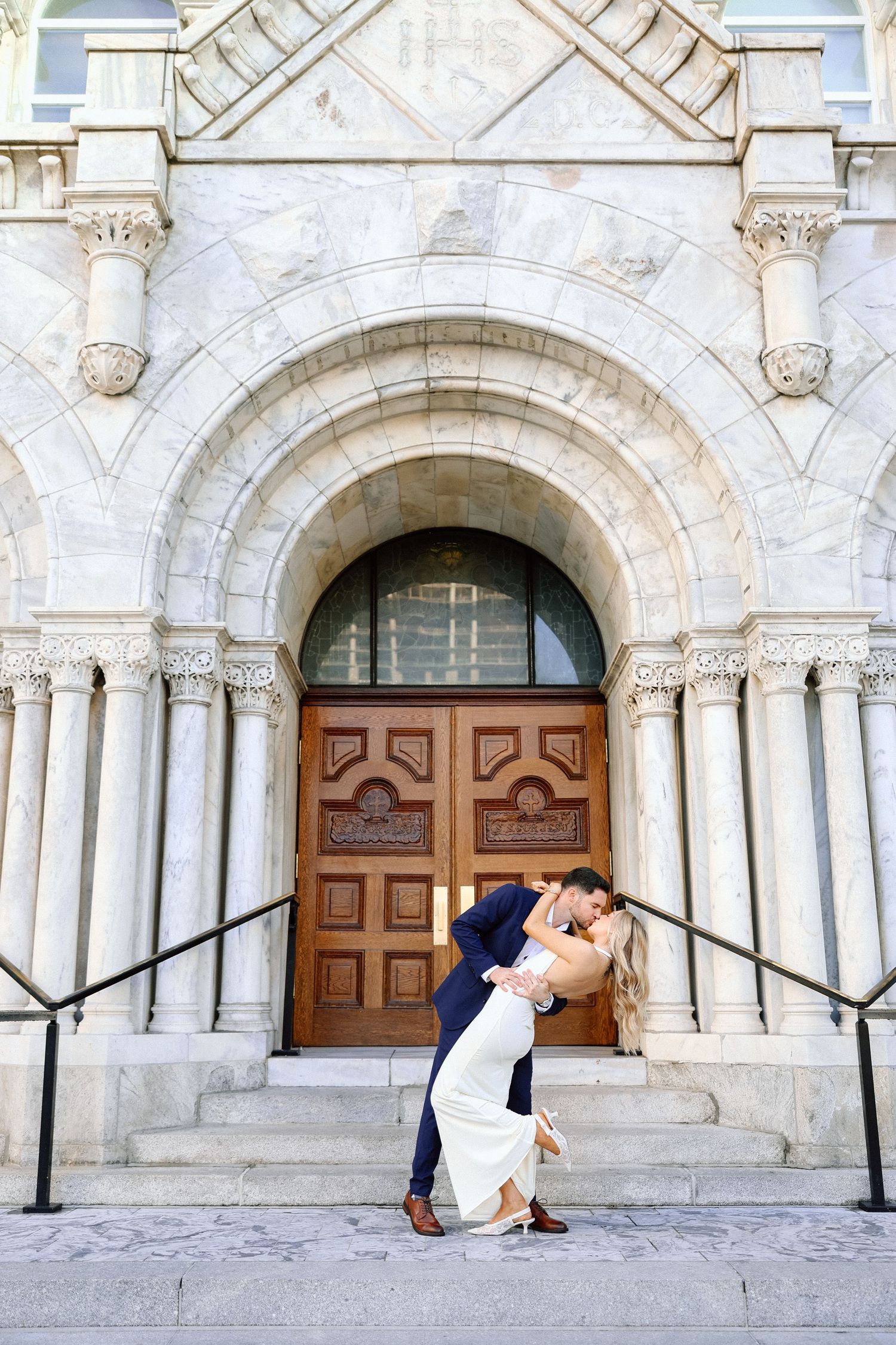 Groom dipping bride in front of ornate wooden doors at a classic stone church entrance.