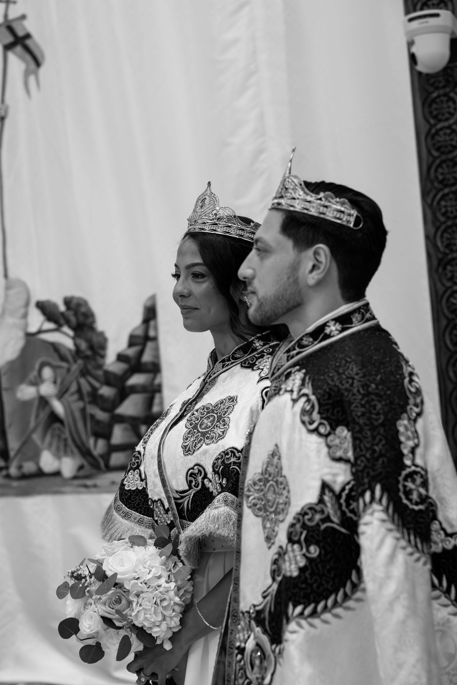 Couple in traditional embroidered wedding attire wearing crowns during Orthodox church ceremony.