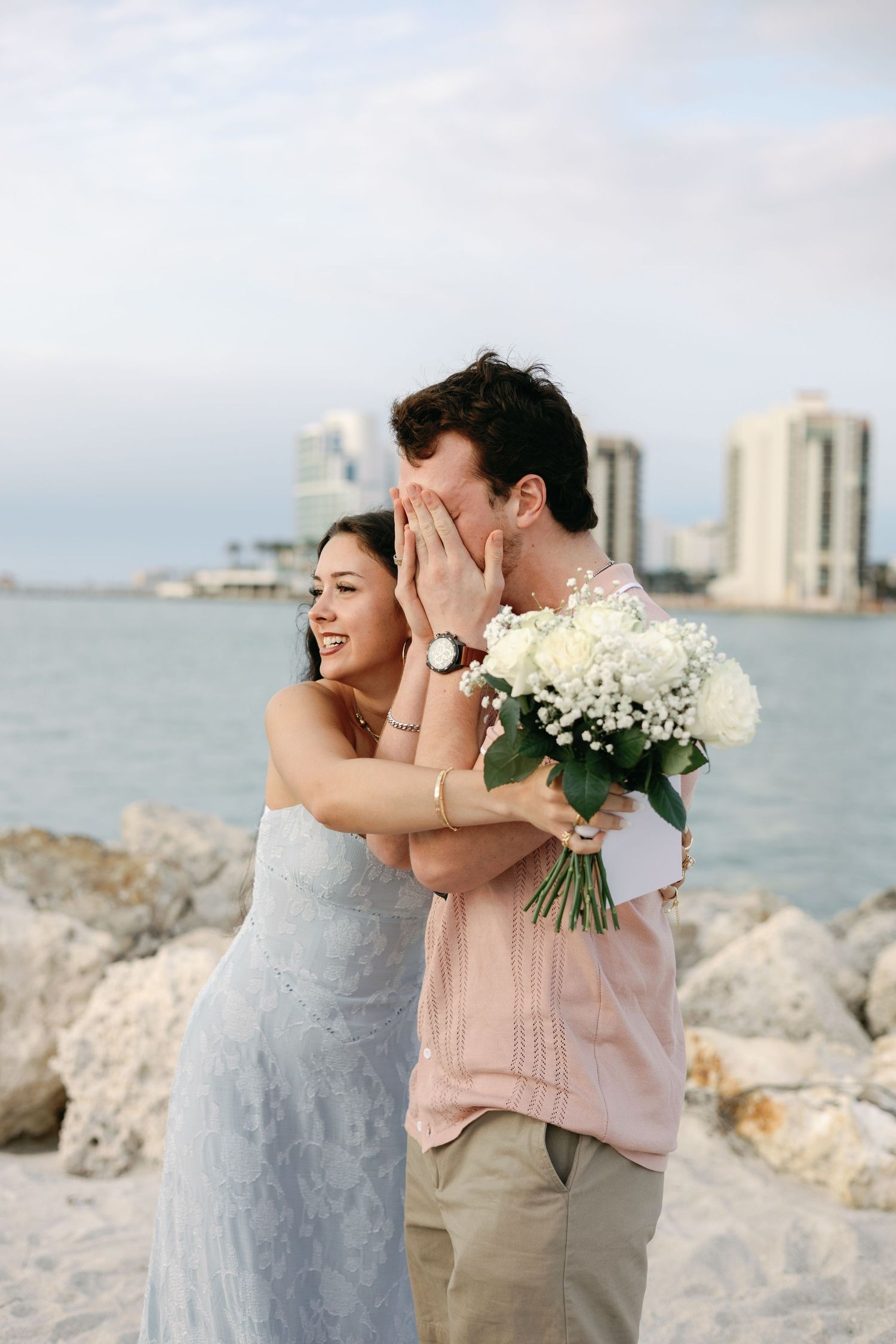 Joyful couple on beach, bride laughing while groom covers face, holding white flower bouquet with city skyline backdrop.
