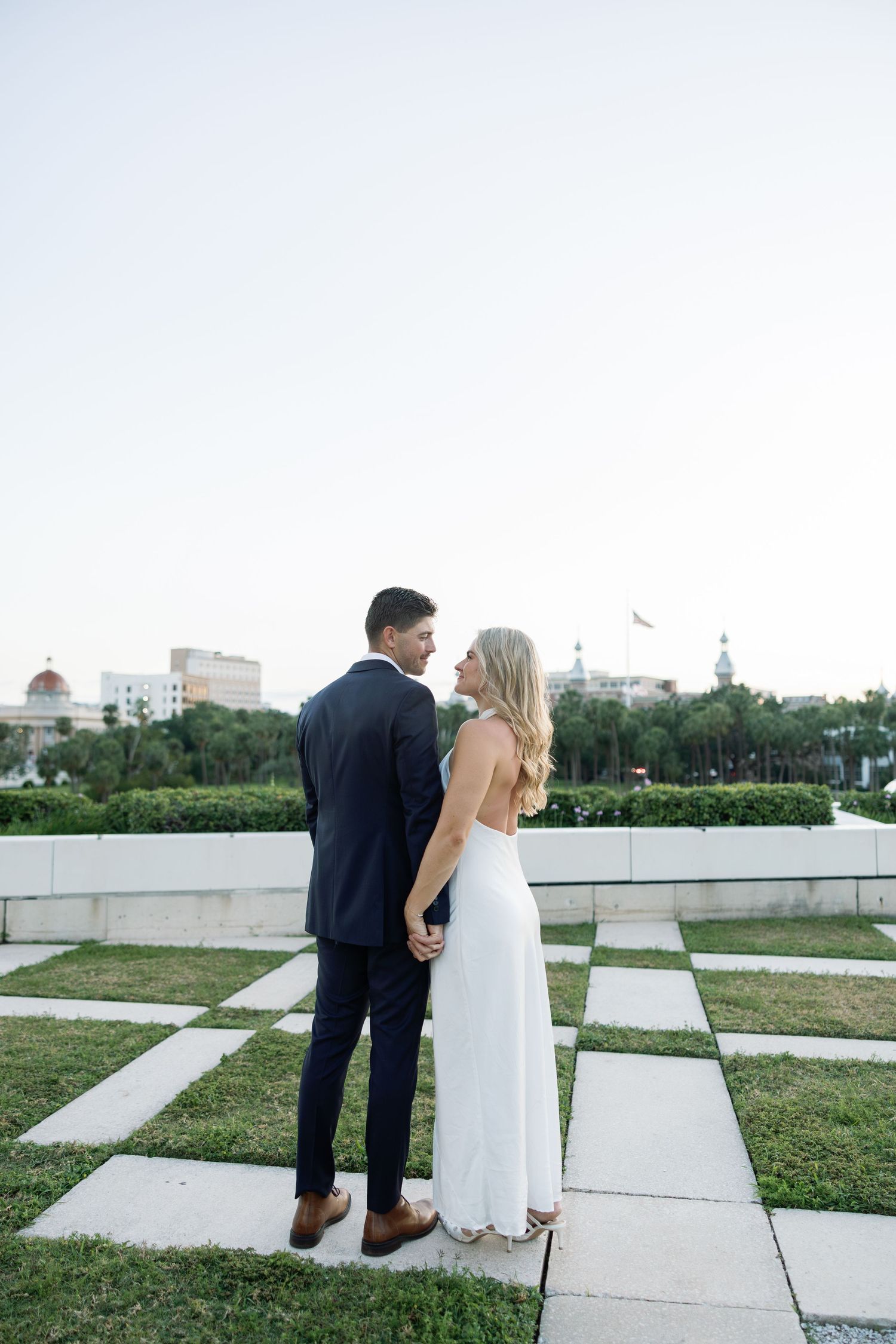 Engaged couple holds hands on checkered plaza with city skyline and palm trees at dusk.