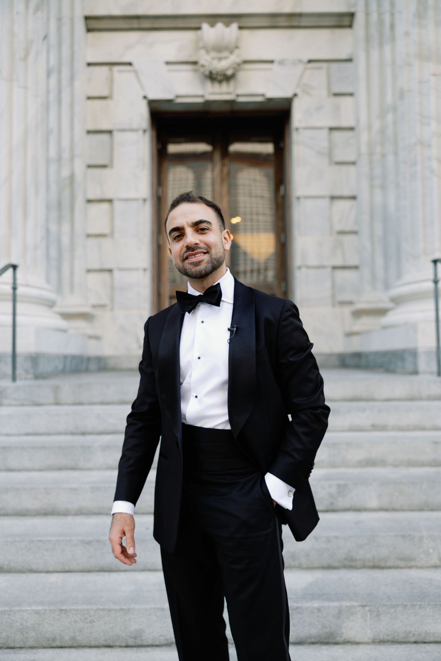 Handsome groom in black tuxedo and bow tie smiling on steps of a grand stone building.