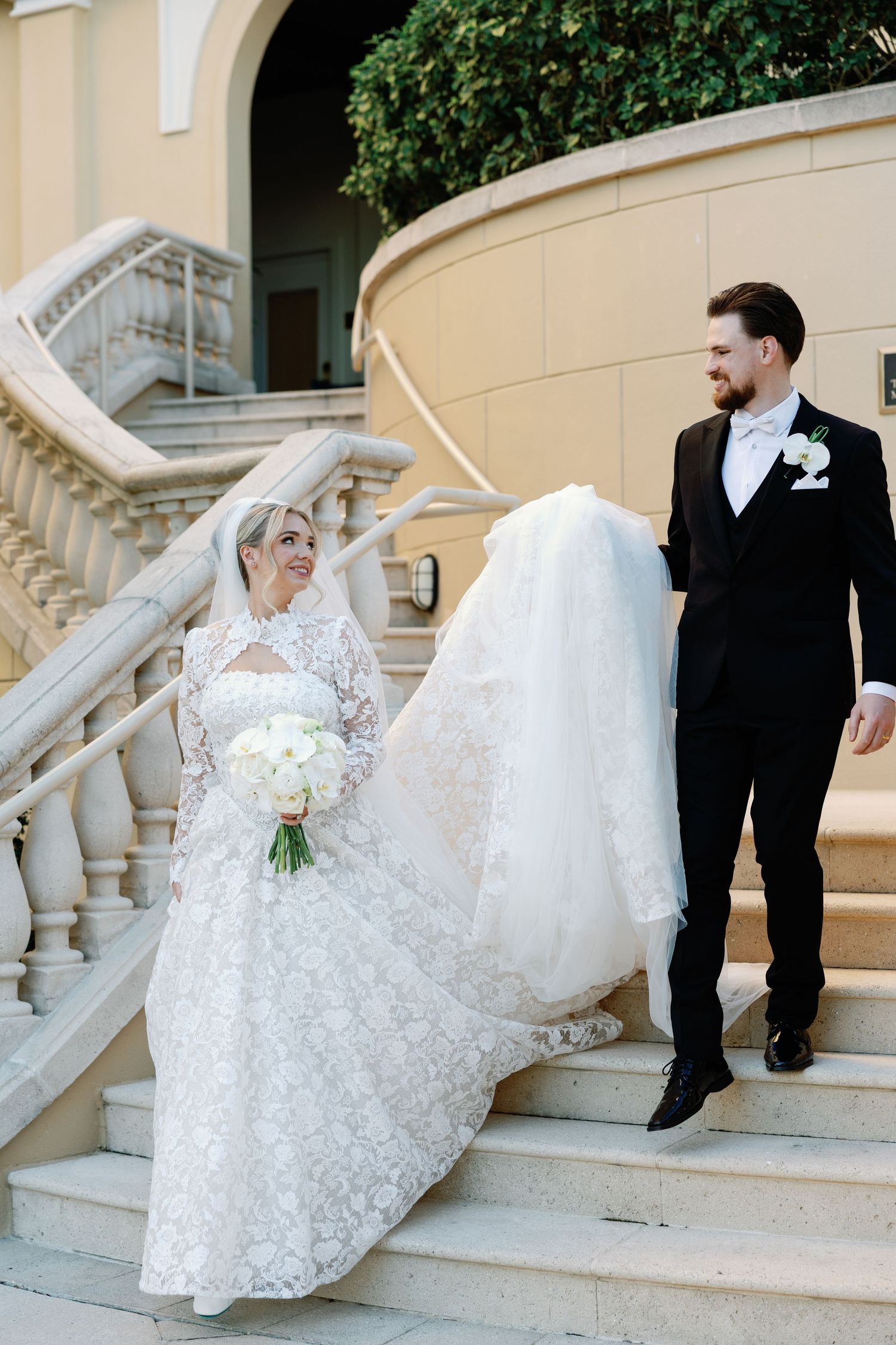 Bride in lace gown and groom in black tuxedo walk down elegant stone staircase at wedding venue.