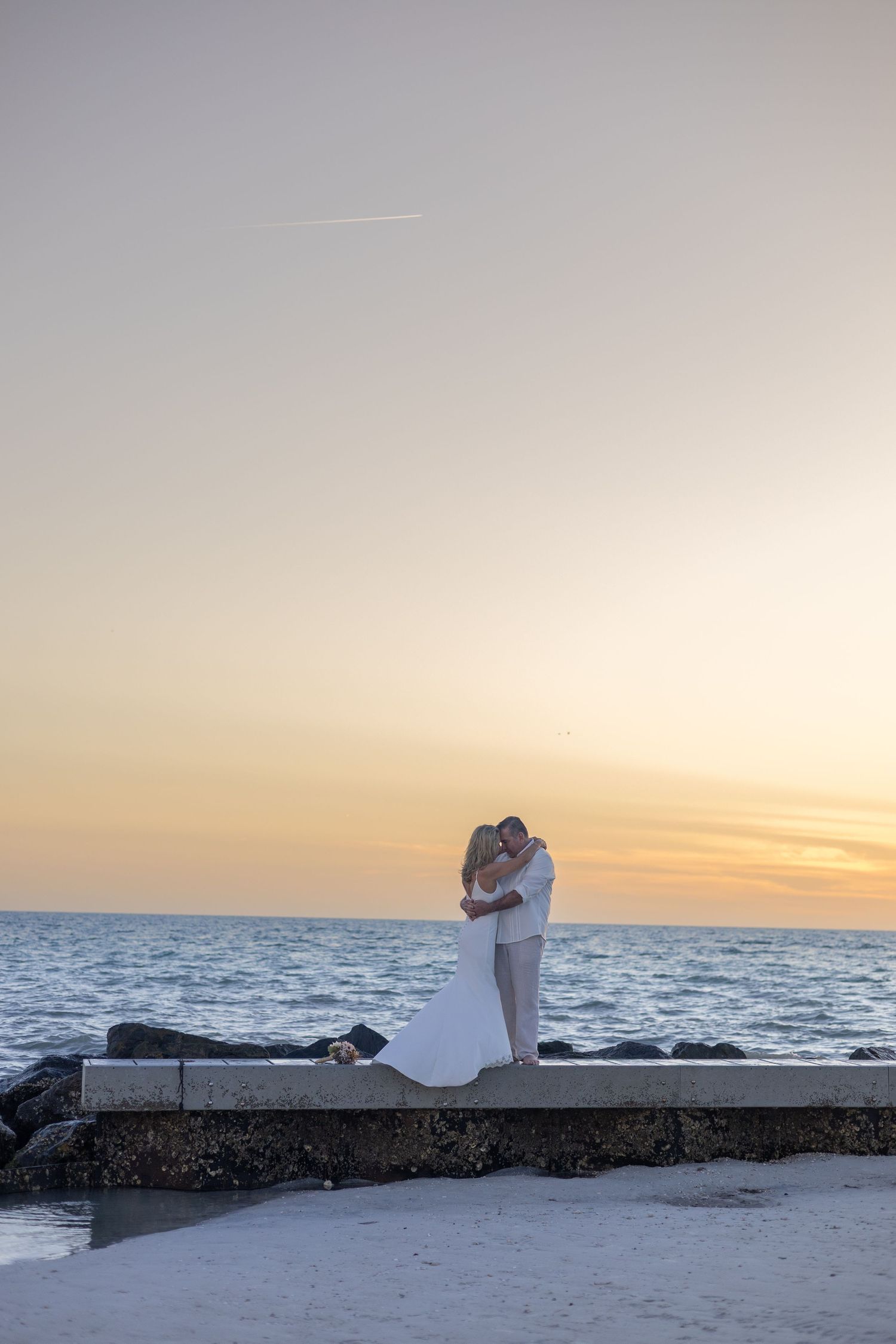 Newlyweds sharing romantic kiss on stone jetty overlooking ocean at stunning golden sunset.