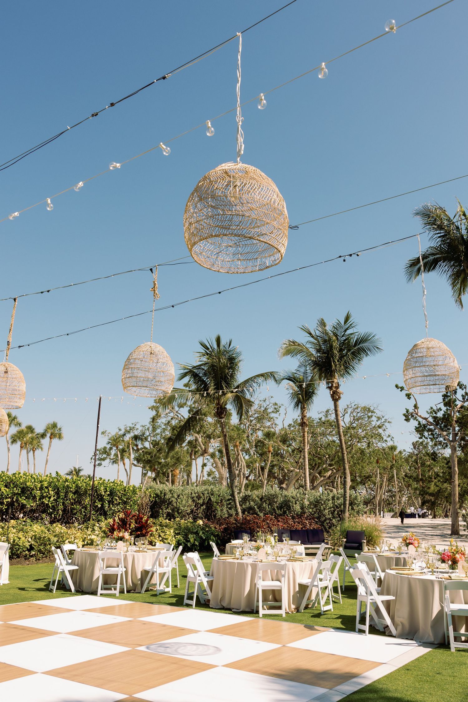 Outdoor beach wedding reception with checkered dance floor, wicker pendant lights, string lights, and palm trees.