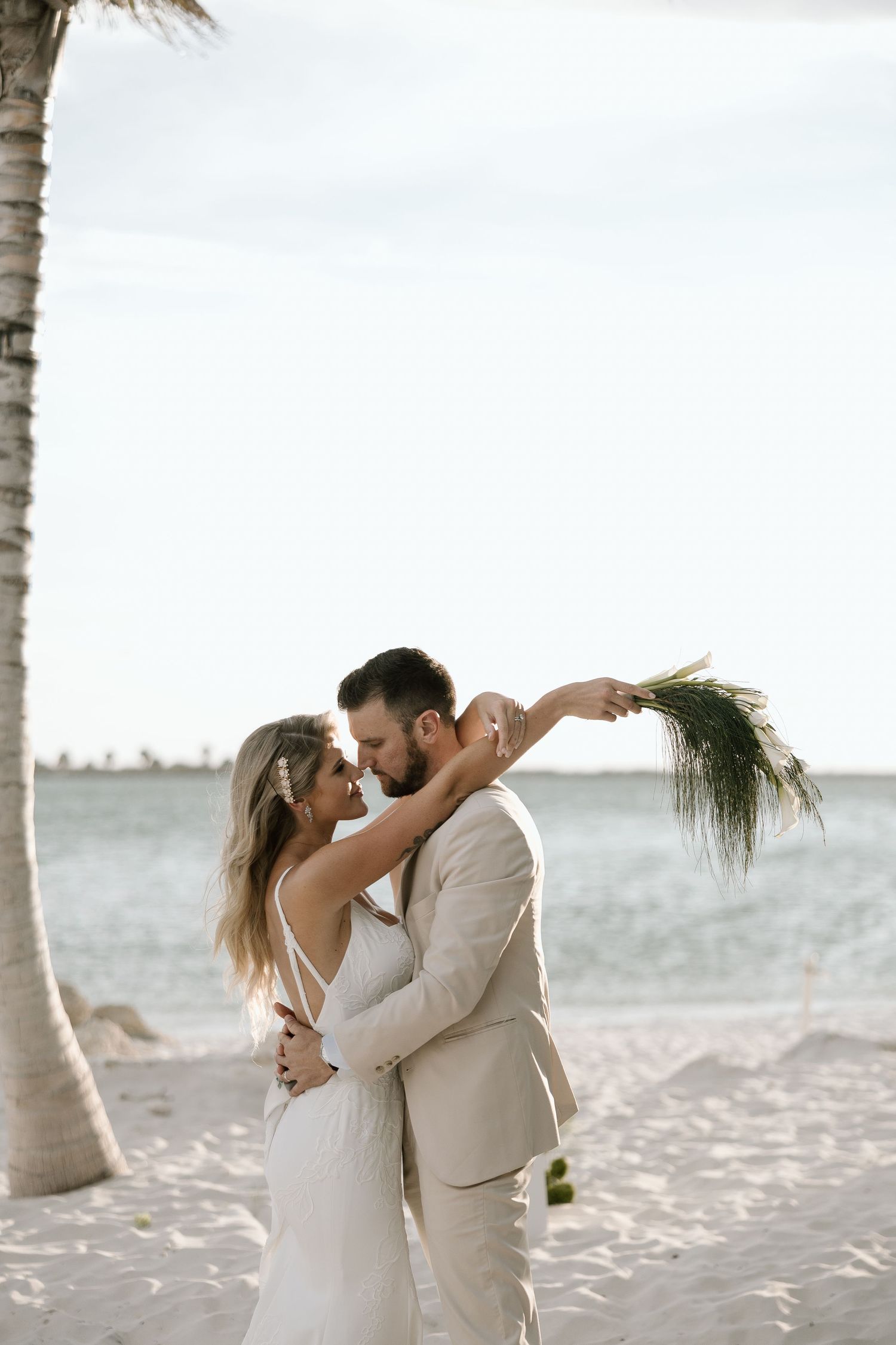 Bride and groom sharing romantic kiss under palm tree on white sand beach, bride holding tropical greenery bouquet.