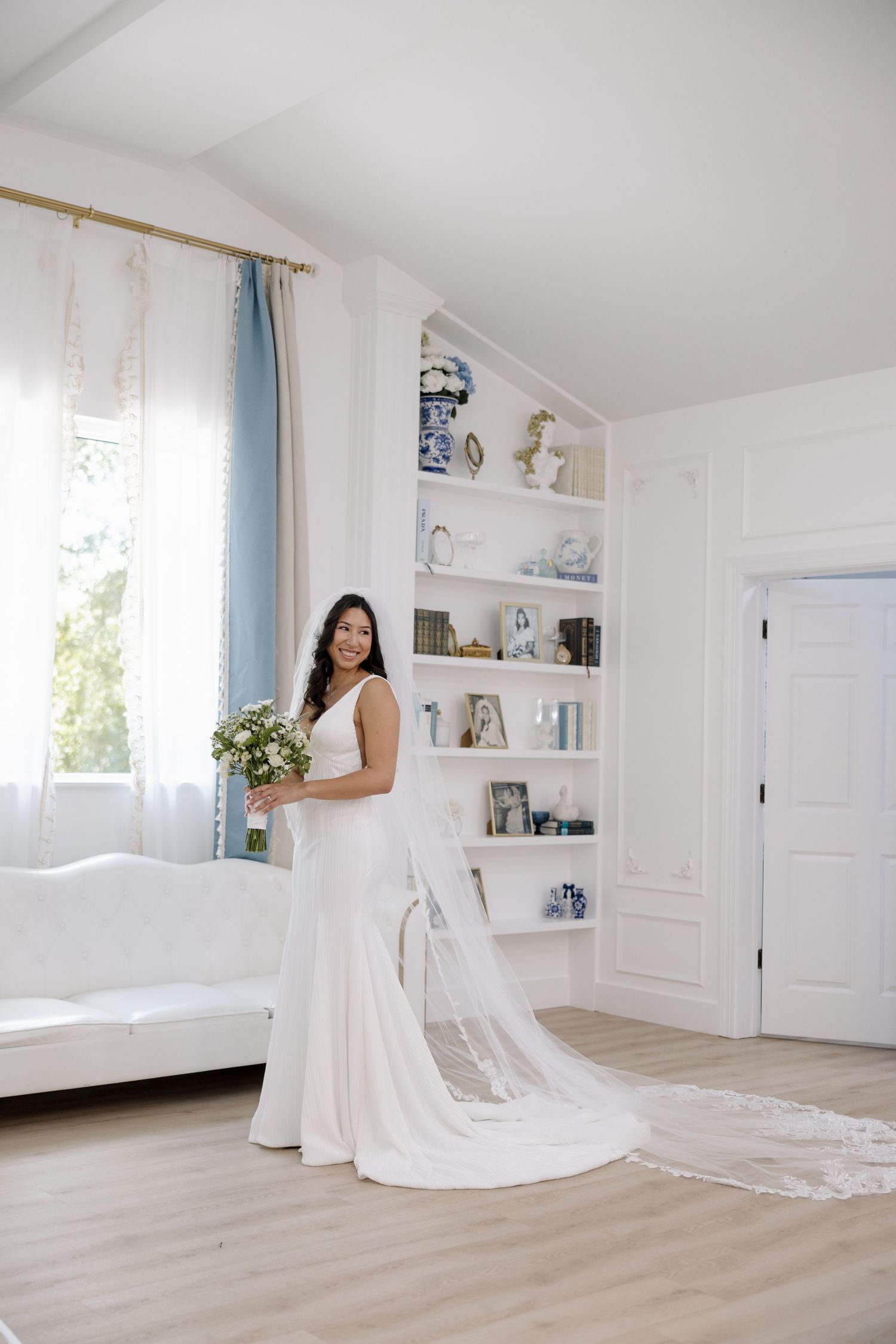 Bride in fitted white lace gown holds bouquet in bright white room with built-in bookshelves.