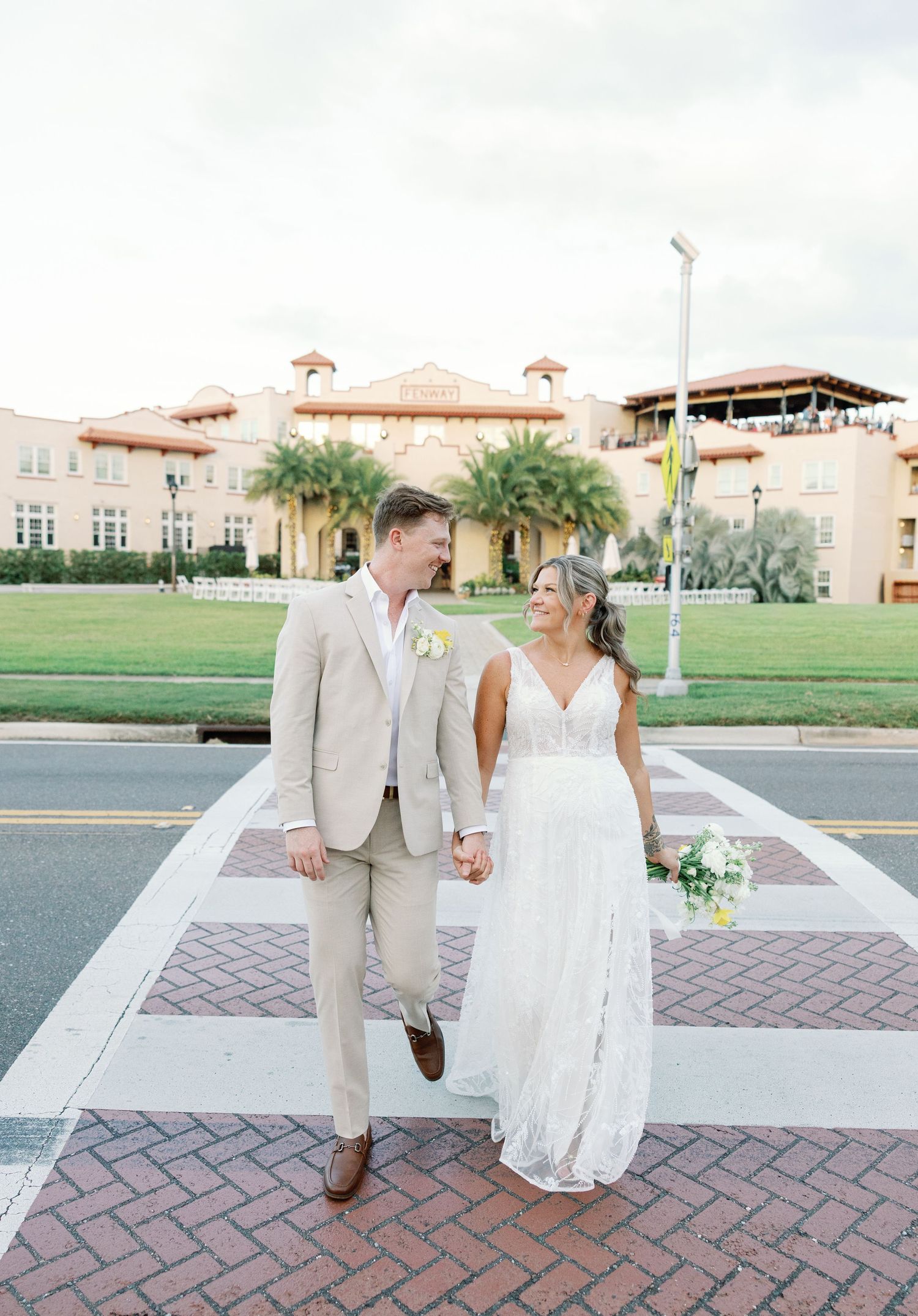 Bride and groom holding hands walking across street with historic Spanish-style wedding venue behind them.