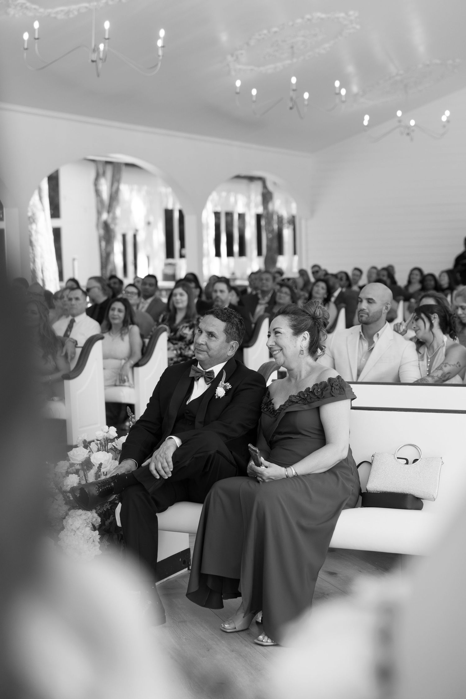Black and white photo of wedding guests smiling and seated in white pews during ceremony.