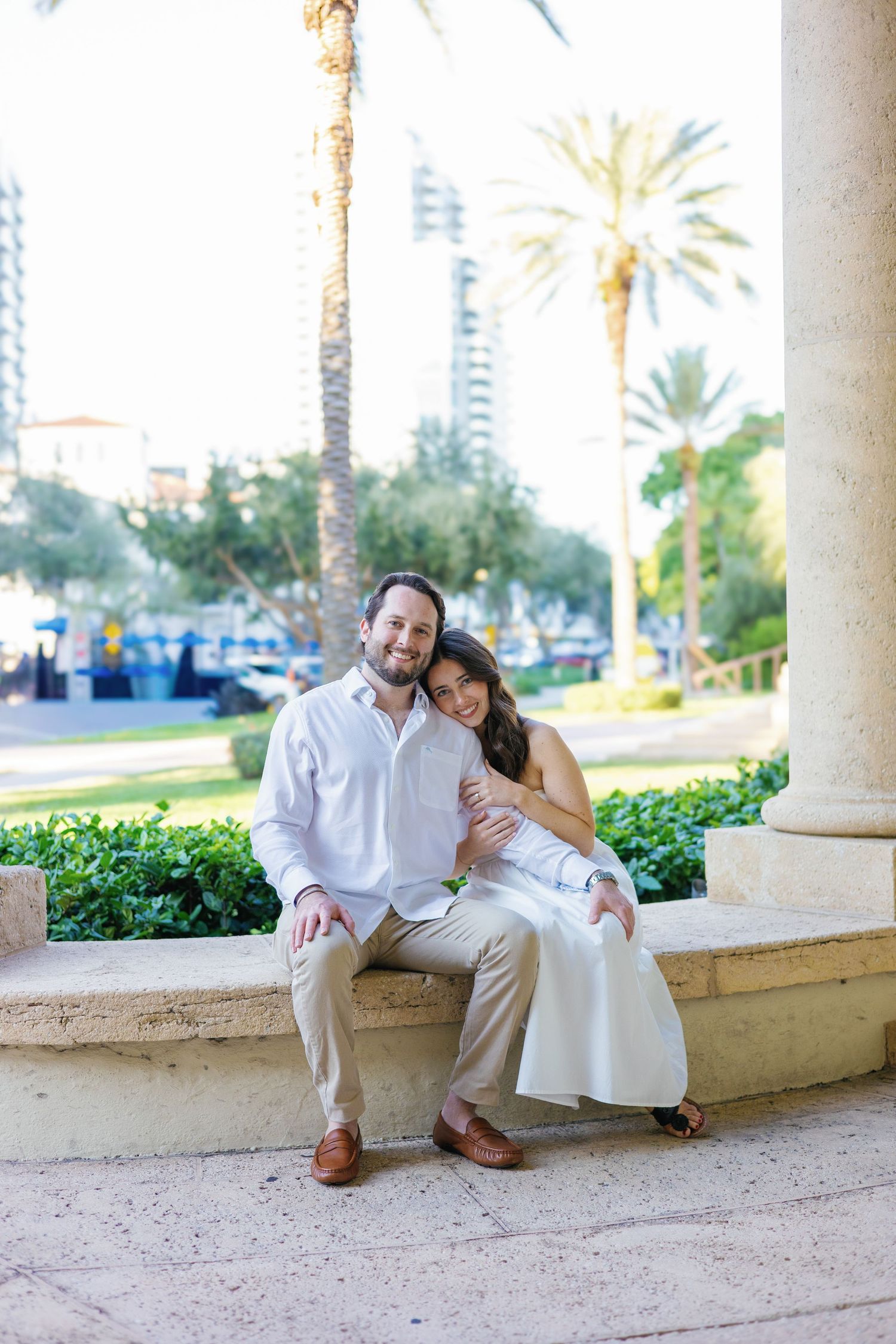 Smiling couple sitting together on stone ledge surrounded by tropical palm trees.
