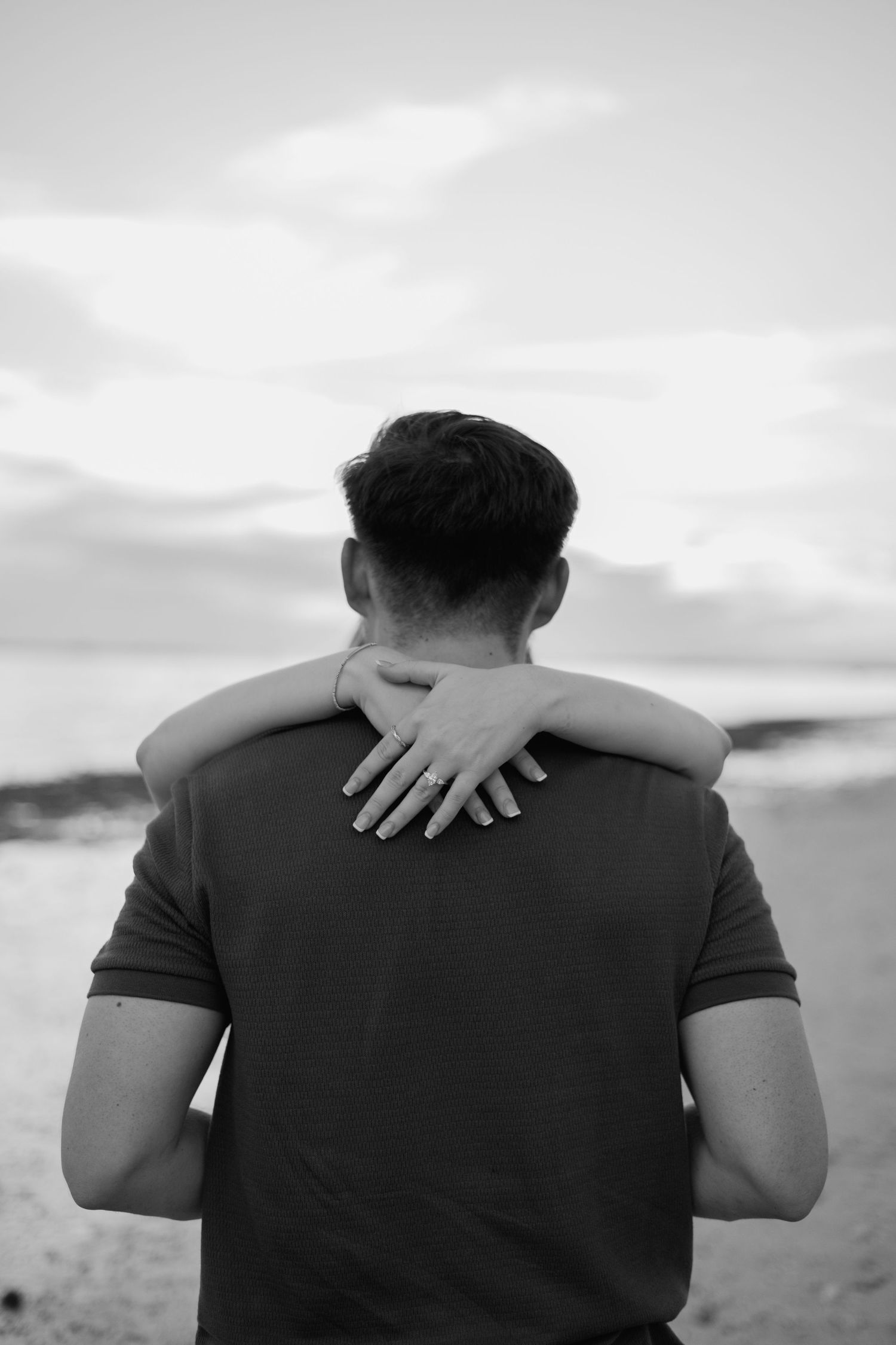 Black and white photo of woman placing hand with engagement ring on man's shoulder at the beach.