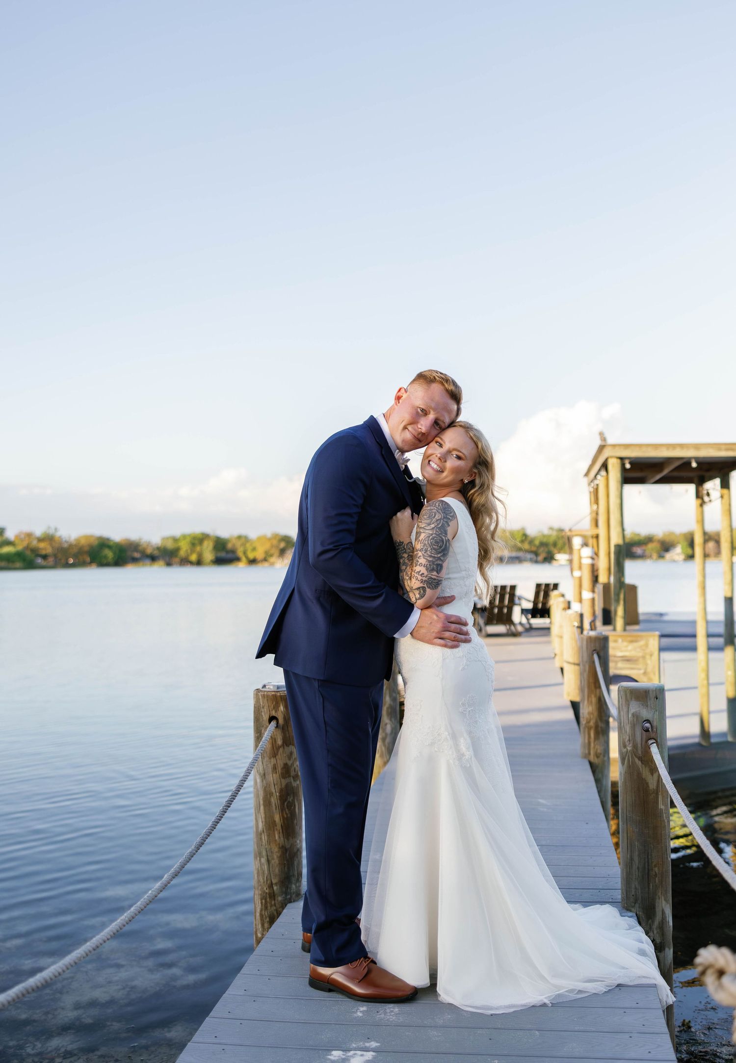 Smiling bride and groom pose together on waterfront dock at sunset after outdoor lakeside wedding ceremony.