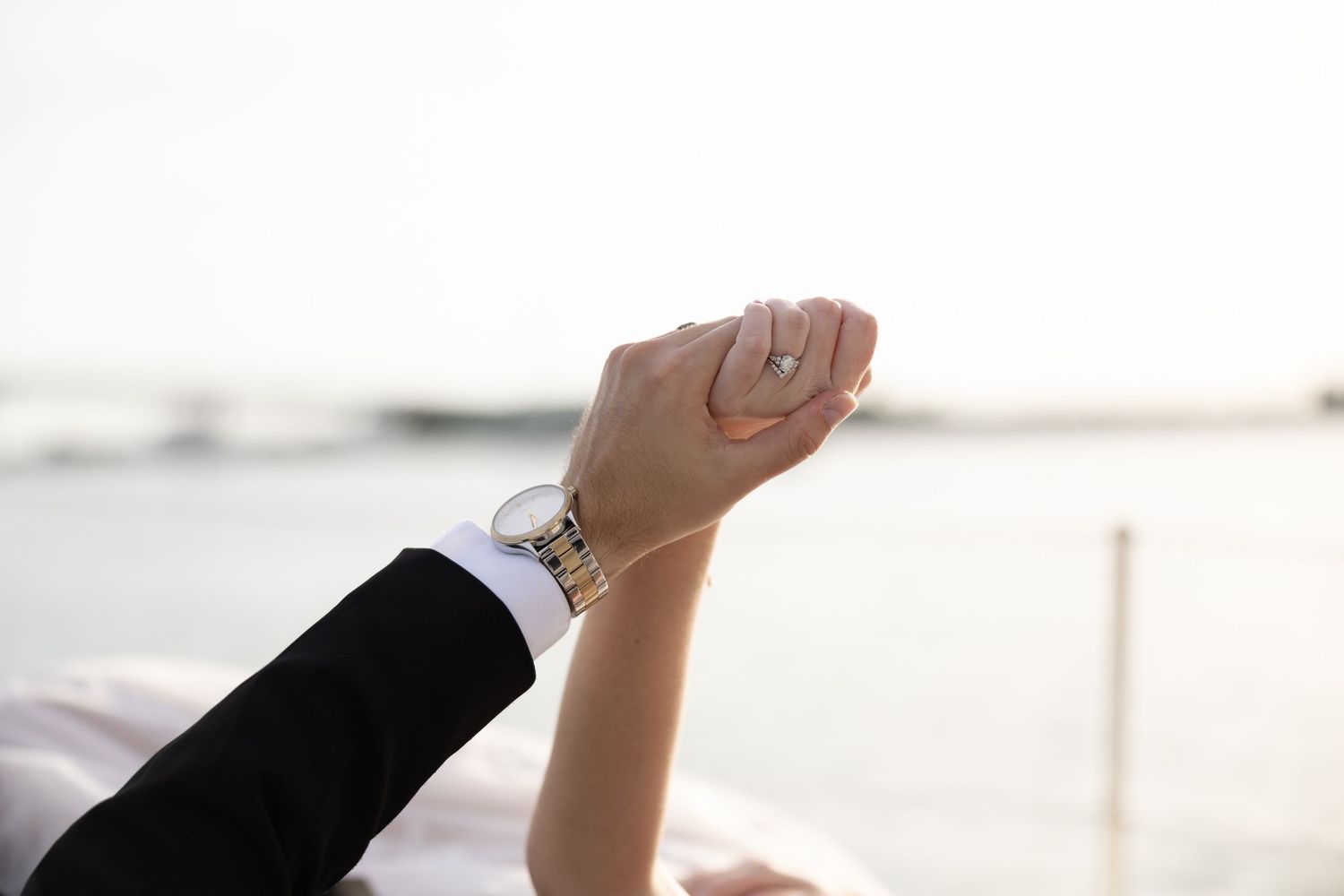 Close-up of bride's hand with diamond ring and gold watch raised against a bright waterfront background.