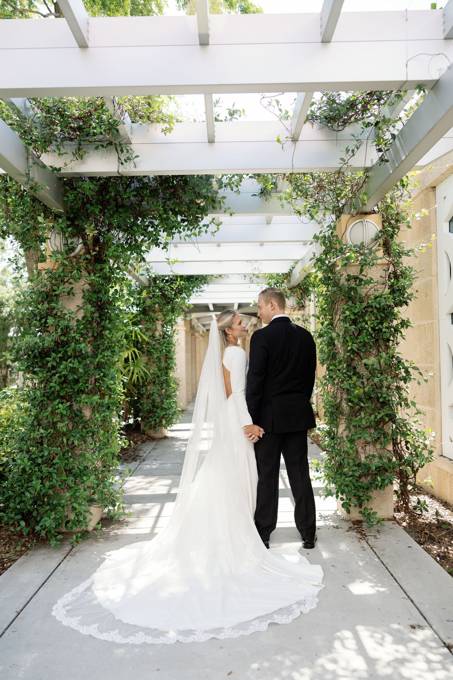Bride and groom share romantic kiss under lush green pergola in elegant outdoor wedding portrait.