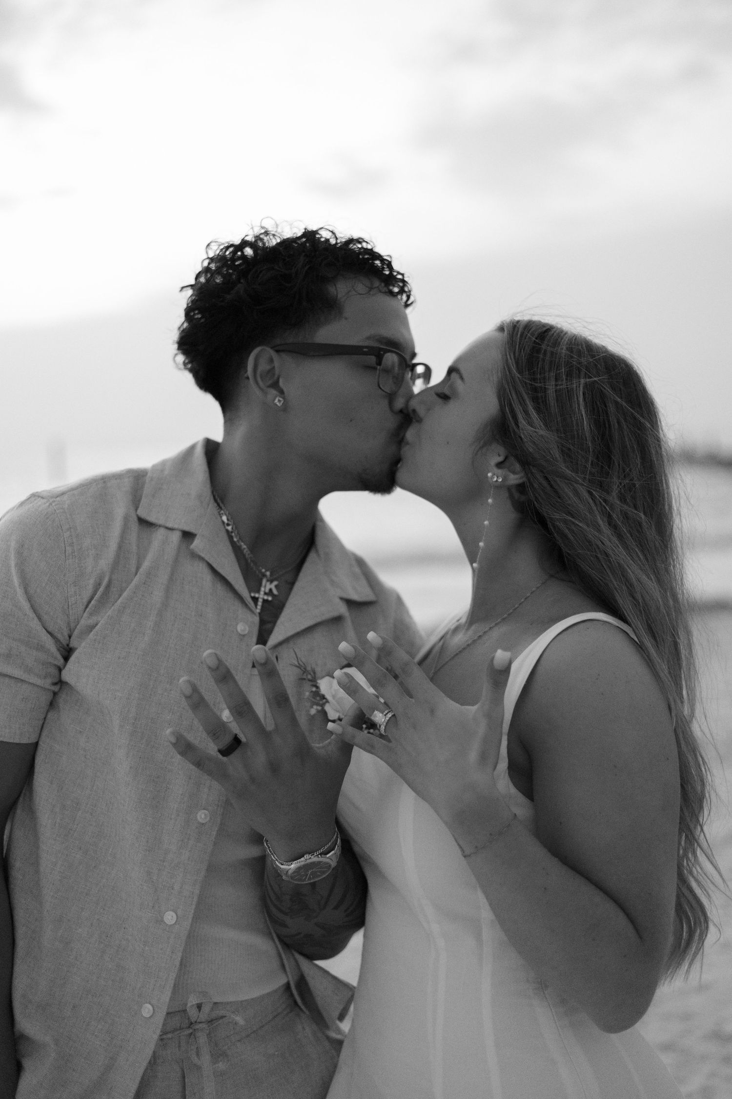 Black and white photo of couple kissing and showing wedding rings on beach.
