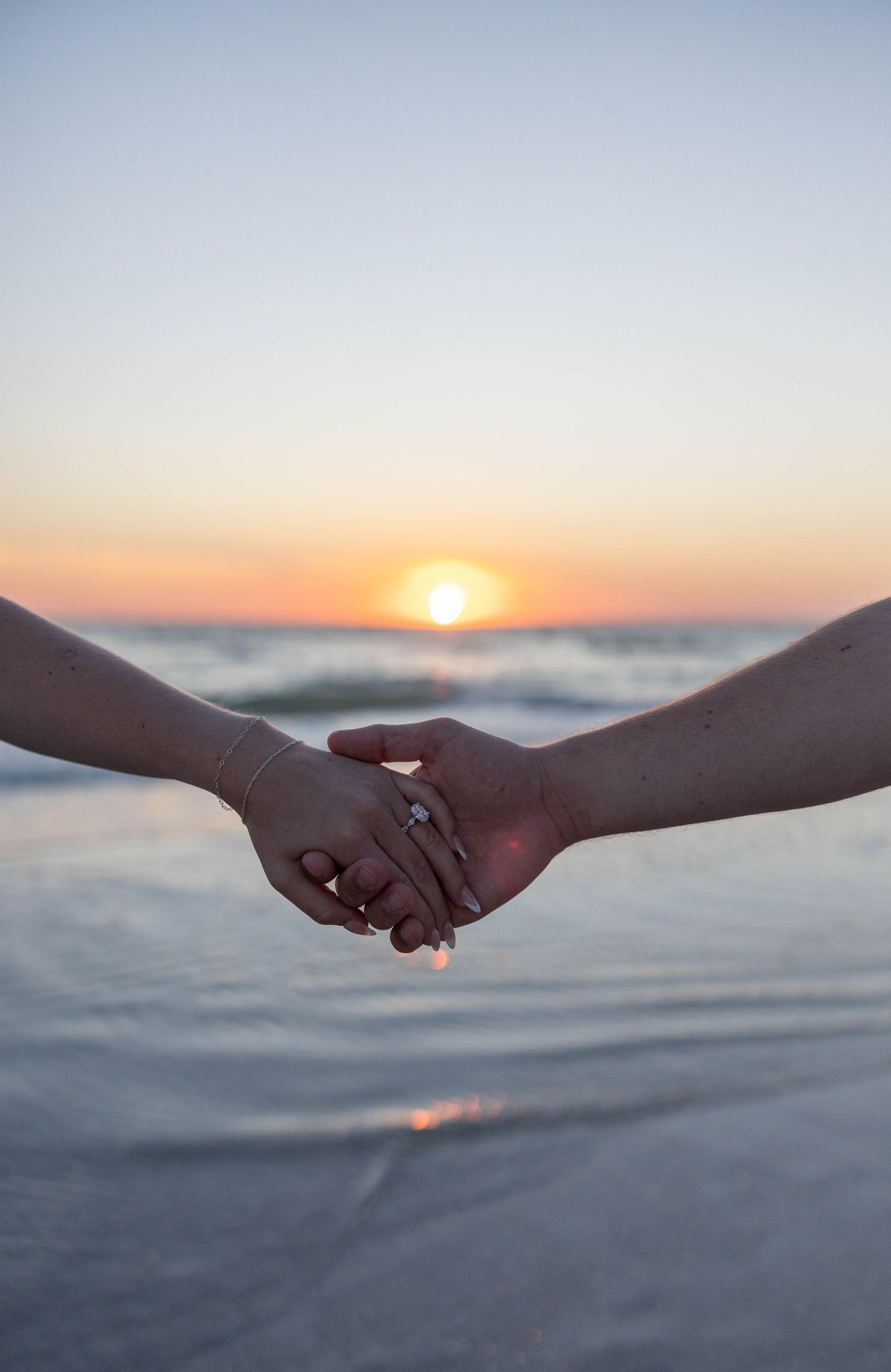 Couple holds hands at sunset on the beach showcasing an engagement ring sparkling in the light.