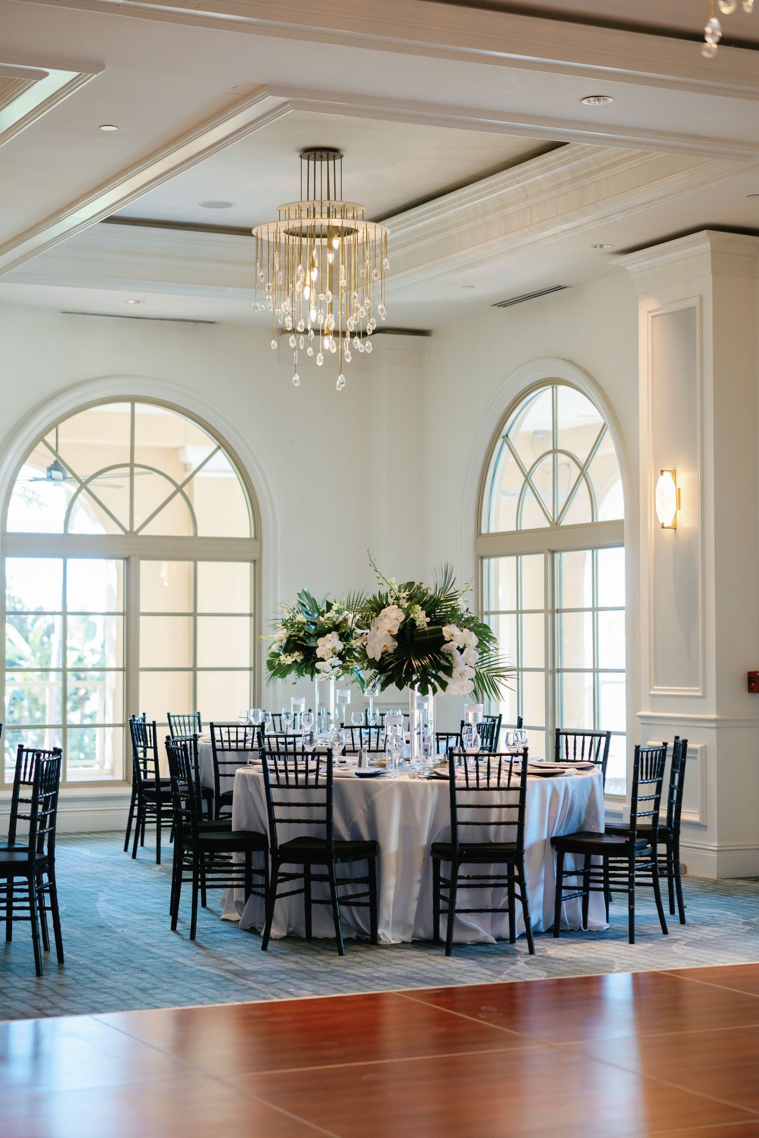 Elegant wedding reception ballroom with black chiavari chairs, floral centerpiece, crystal chandelier, and arched windows.