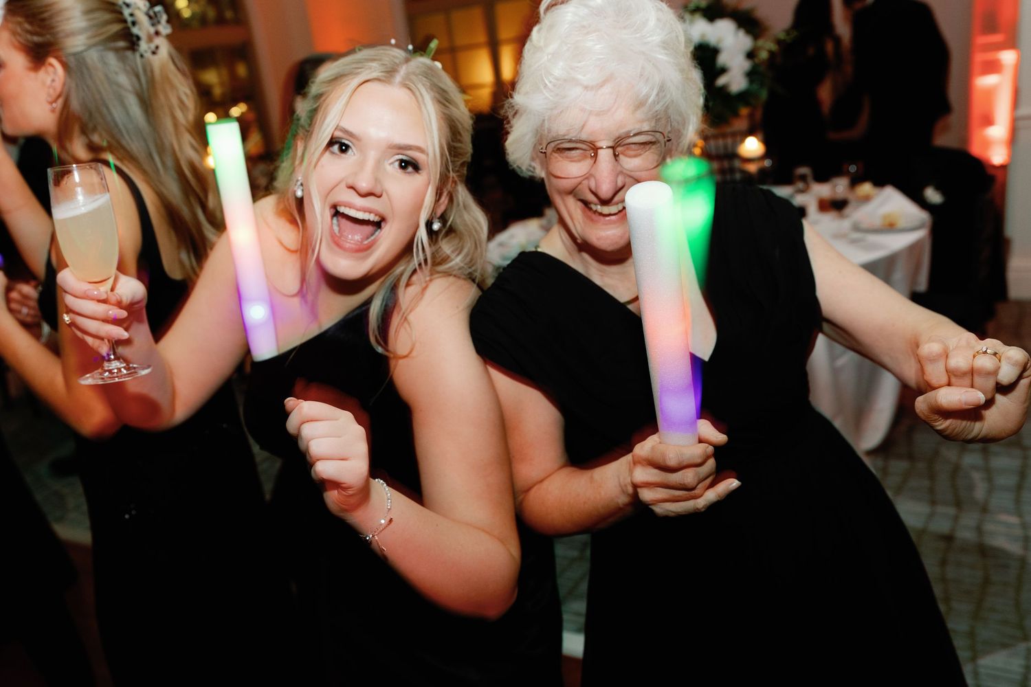 Two women in black dresses dancing joyfully at a wedding reception, holding colorful LED glow sticks.