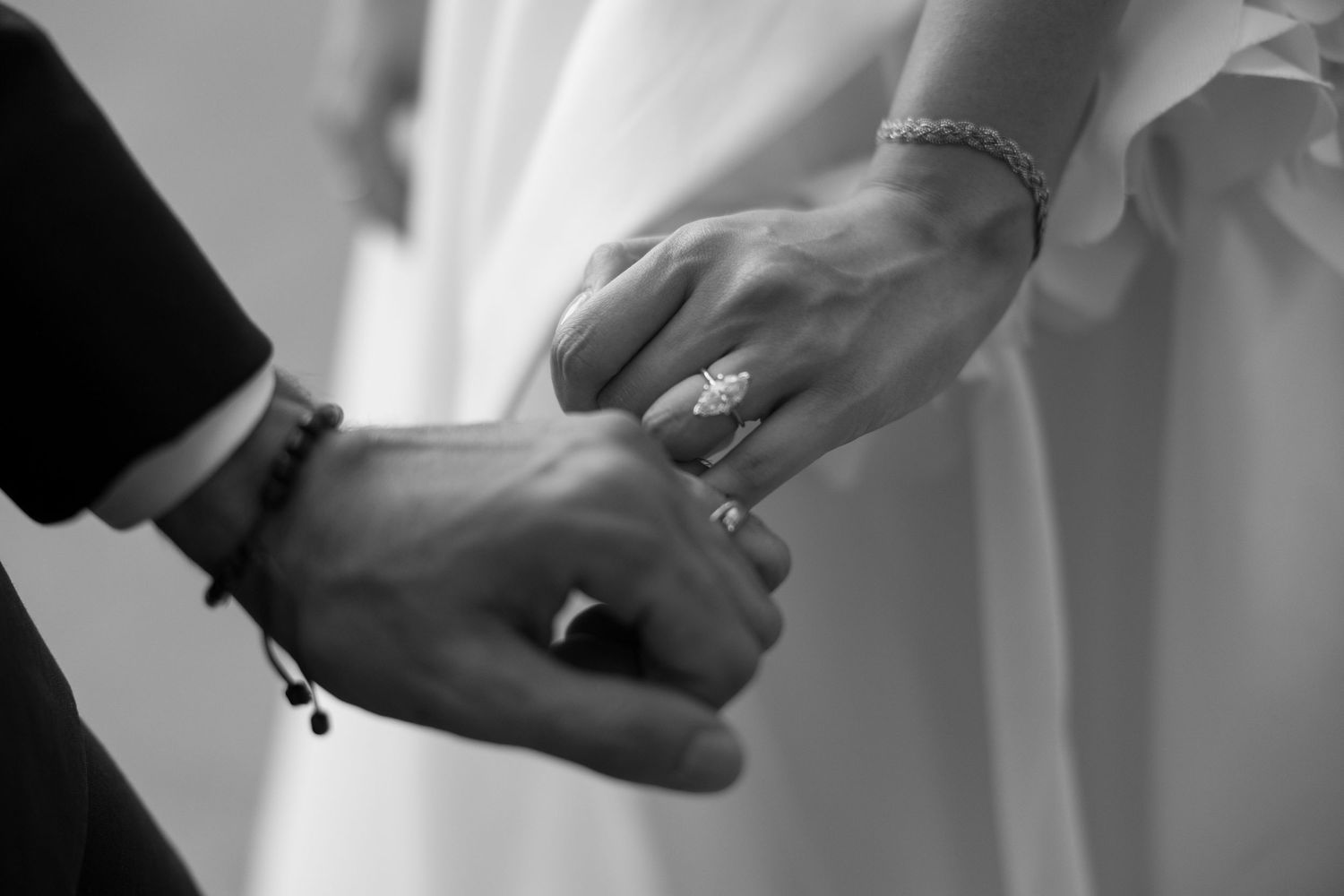 Black and white close-up of bride and groom holding hands showing diamond engagement ring and bracelet.