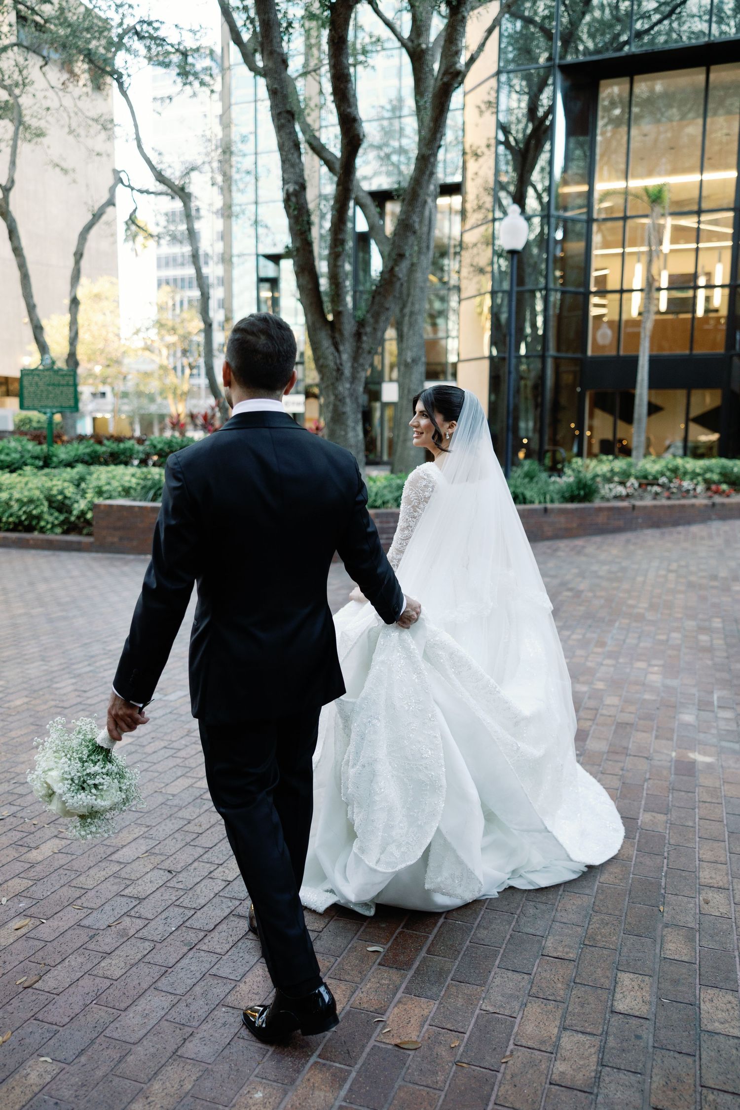 Bride and groom walk hand-in-hand through an urban courtyard, bride in a lace gown with long veil and train.