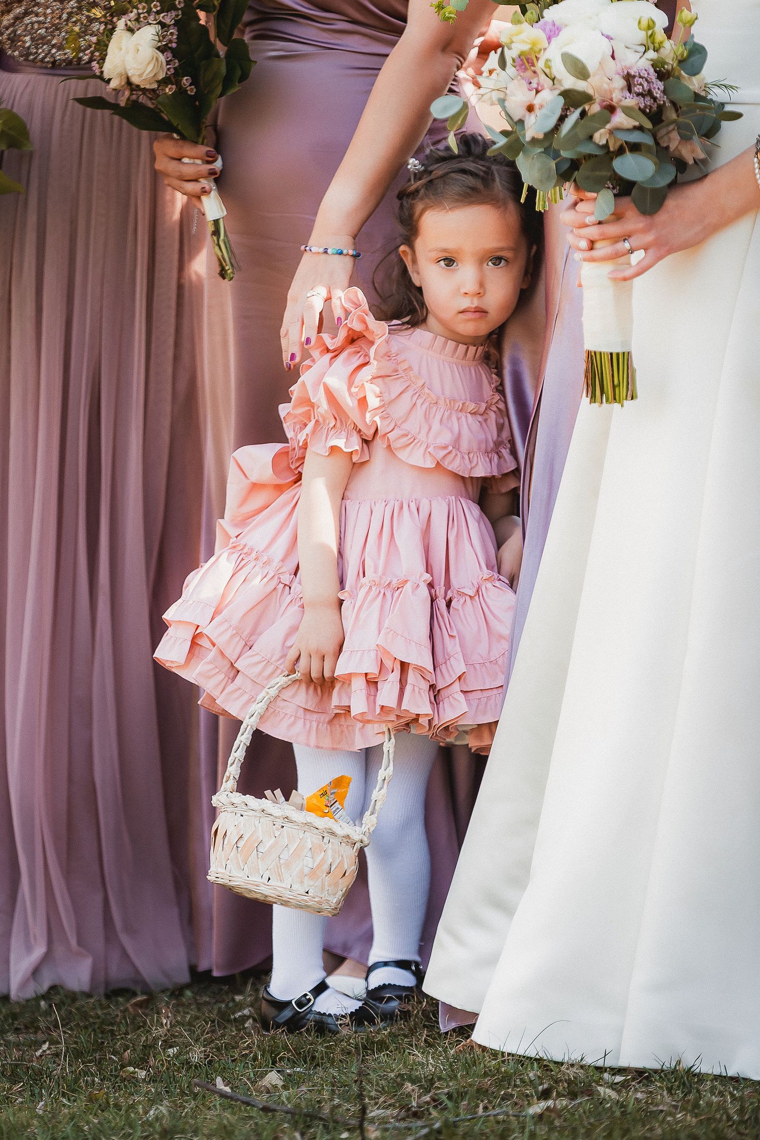 Adorable flower girl in a pink ruffled dress holding a basket standing between bridesmaids.