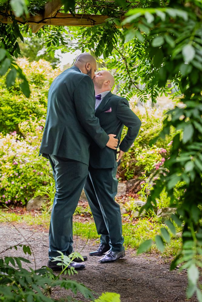 Two grooms in teal suits sharing a kiss beneath a leafy garden archway surrounded by greenery.