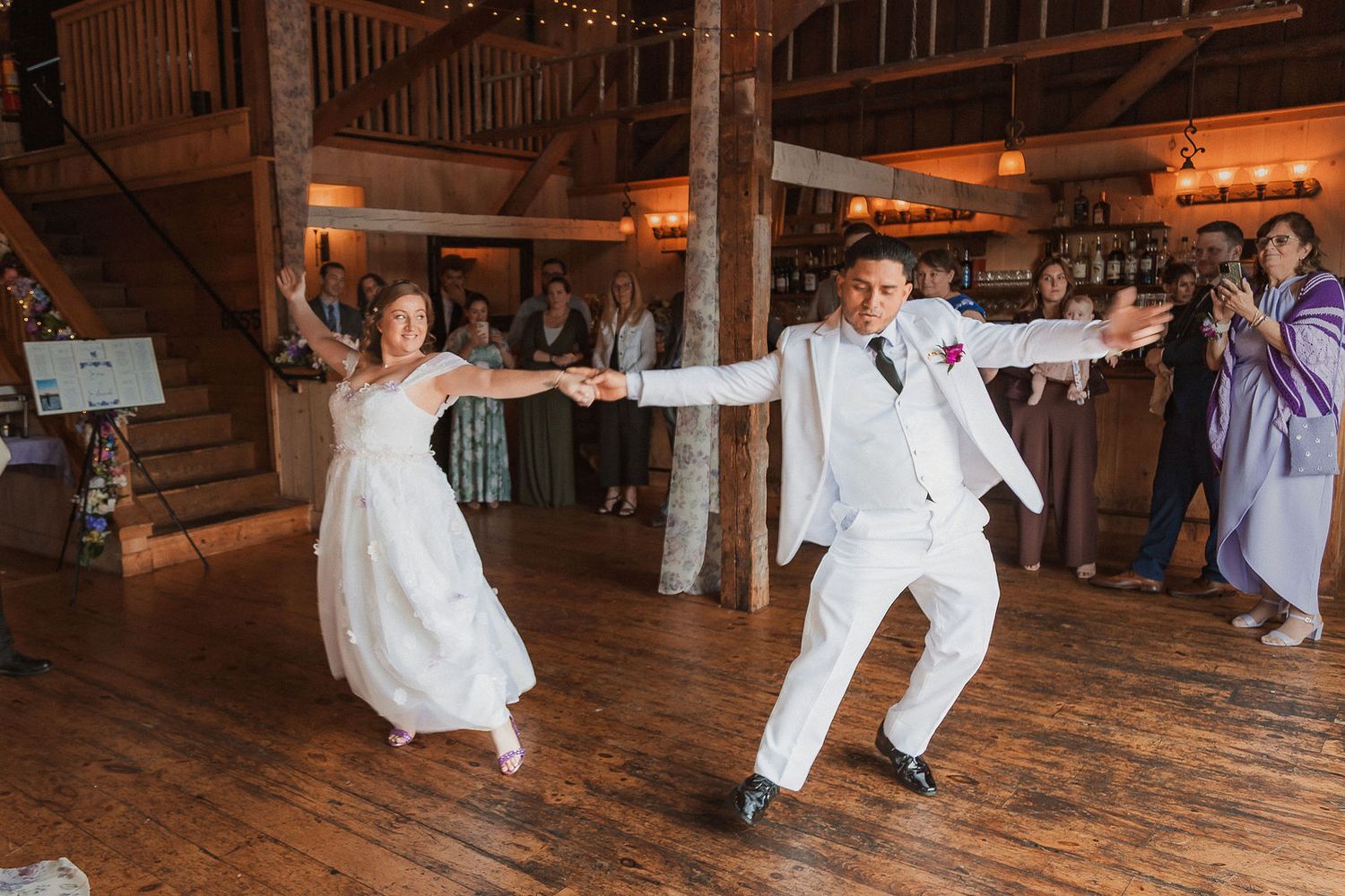 Bride and groom performing energetic first dance in rustic barn wedding venue with warm string lights.