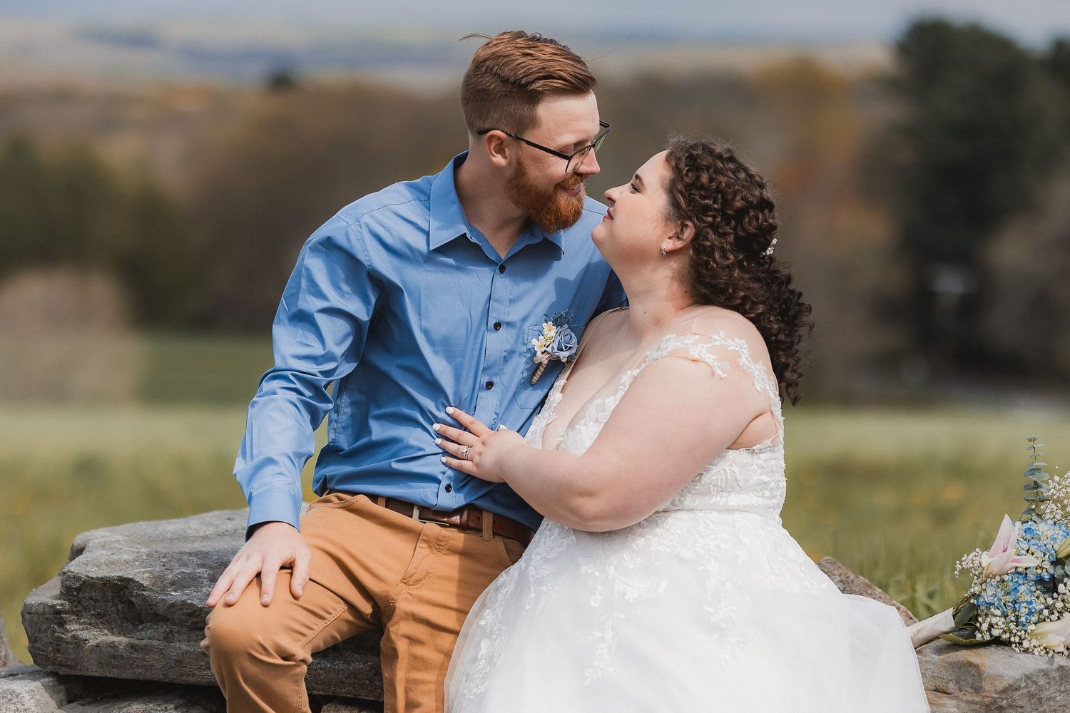 Bride in lace gown and groom in blue shirt share a romantic nose-to-nose moment on a stone wall.