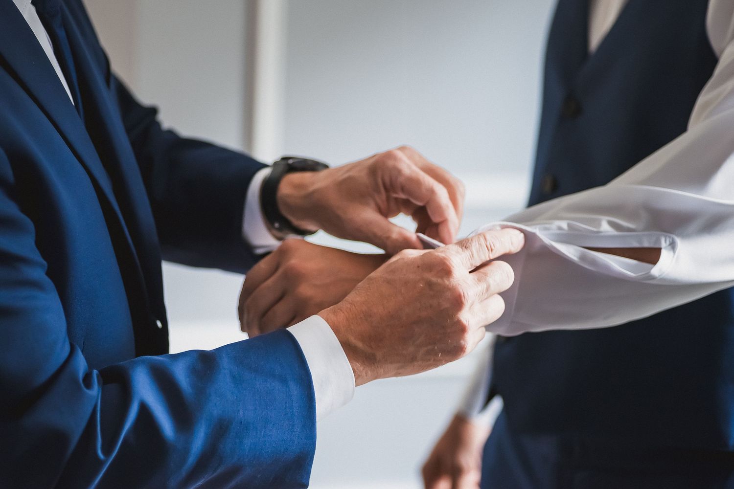 Man in navy suit fastening cufflinks on groom's white dress shirt.