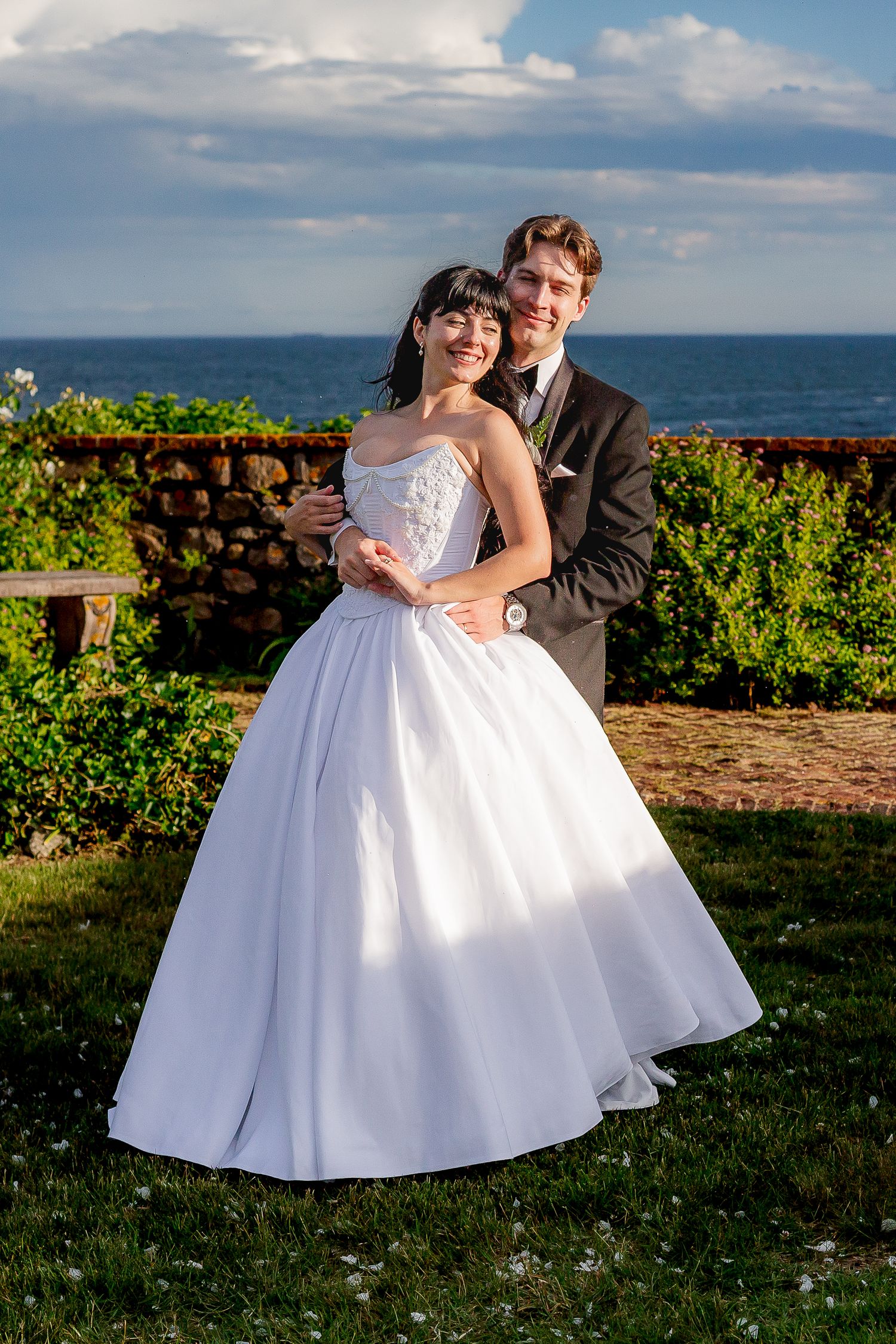Smiling bride in strapless ball gown embraced by groom in dark suit with ocean backdrop.