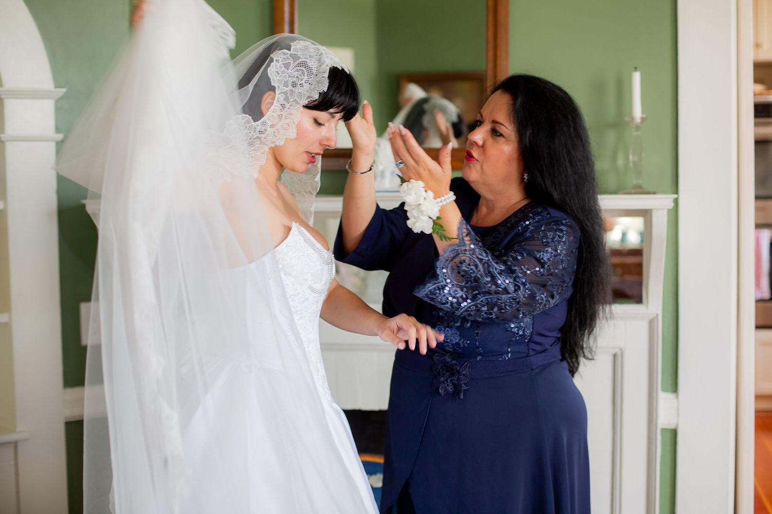 Mother helping bride adjust wedding veil in room with green walls before ceremony.