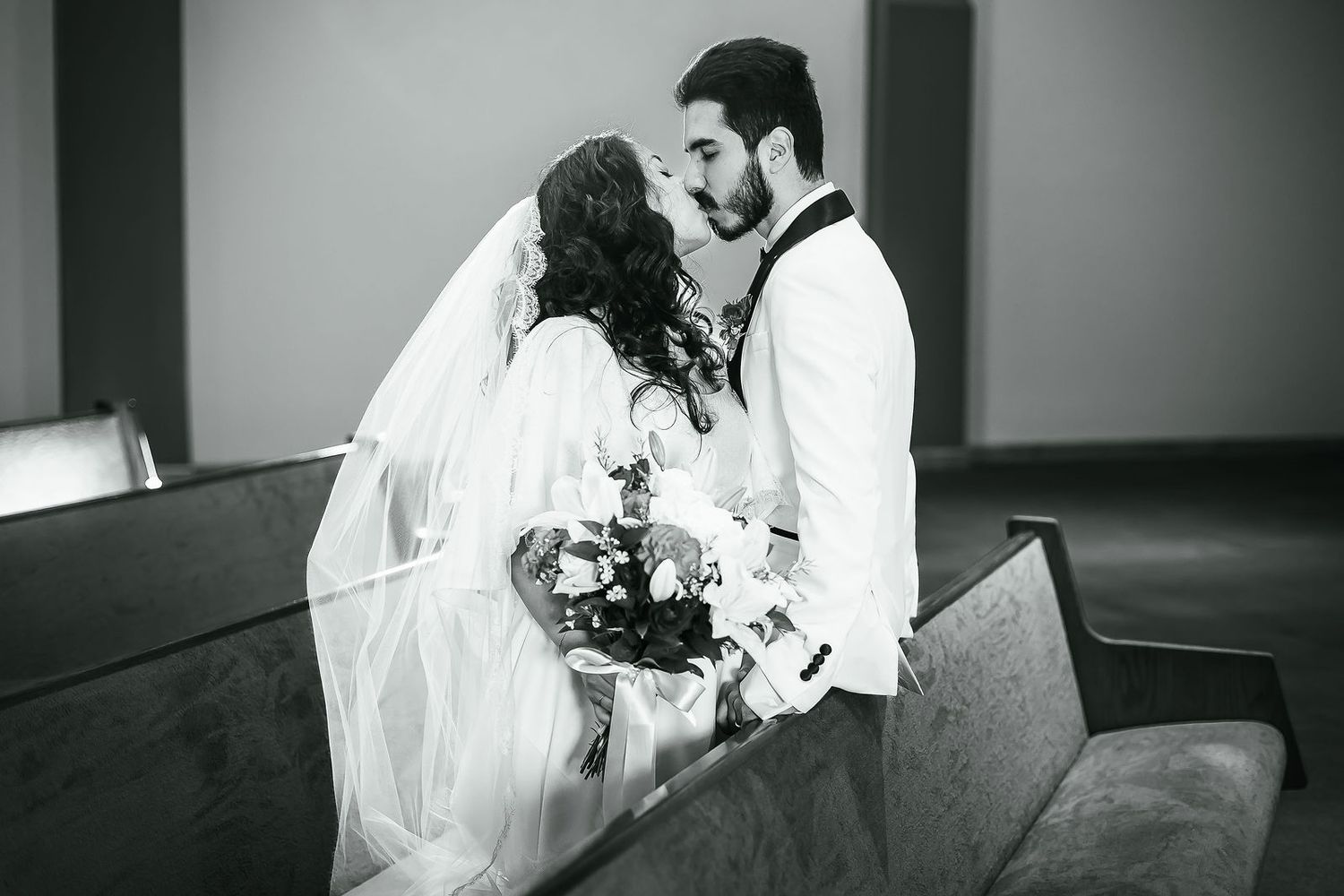 Black and white photo of bride and groom sharing tender kiss in church pew with bridal bouquet.