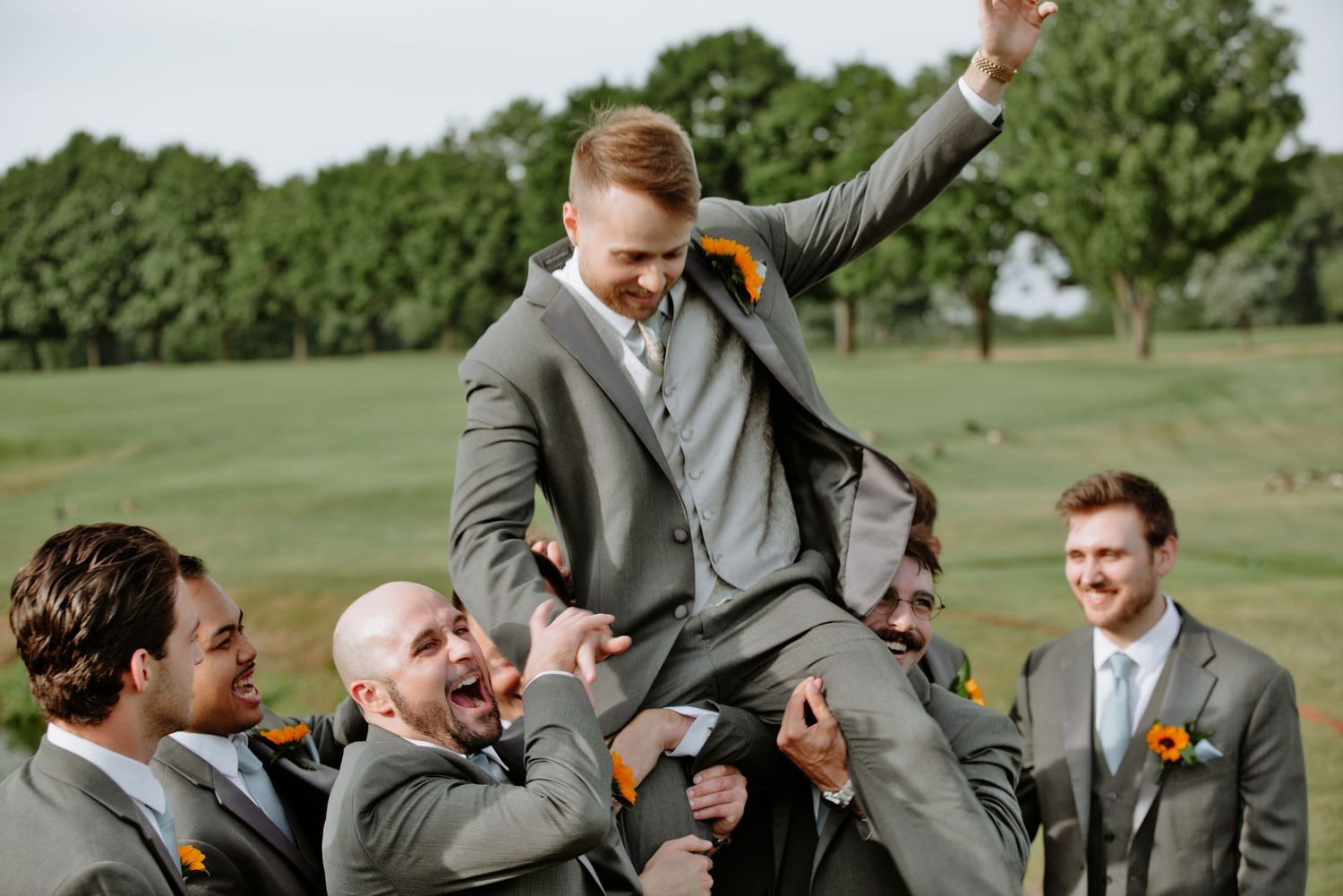 Groomsmen in gray suits lifting groom celebrating outdoors on green lawn with trees behind.