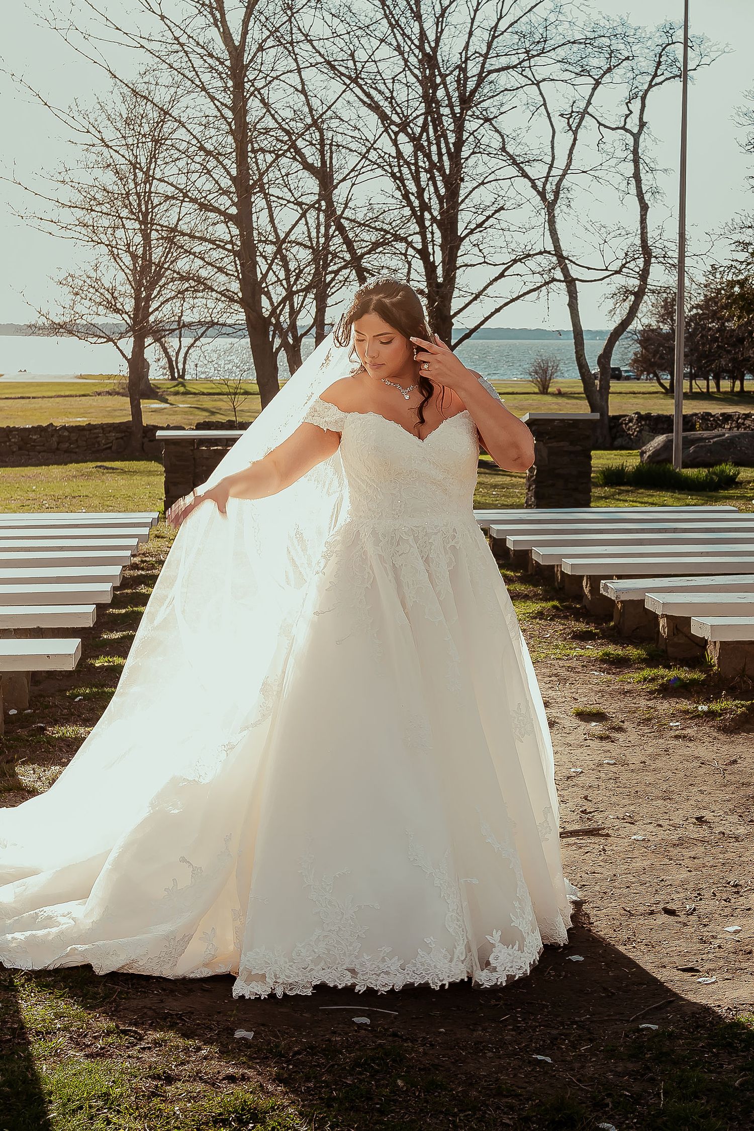 Bride in an off-shoulder lace ball gown with veil posing outdoors near a waterfront.