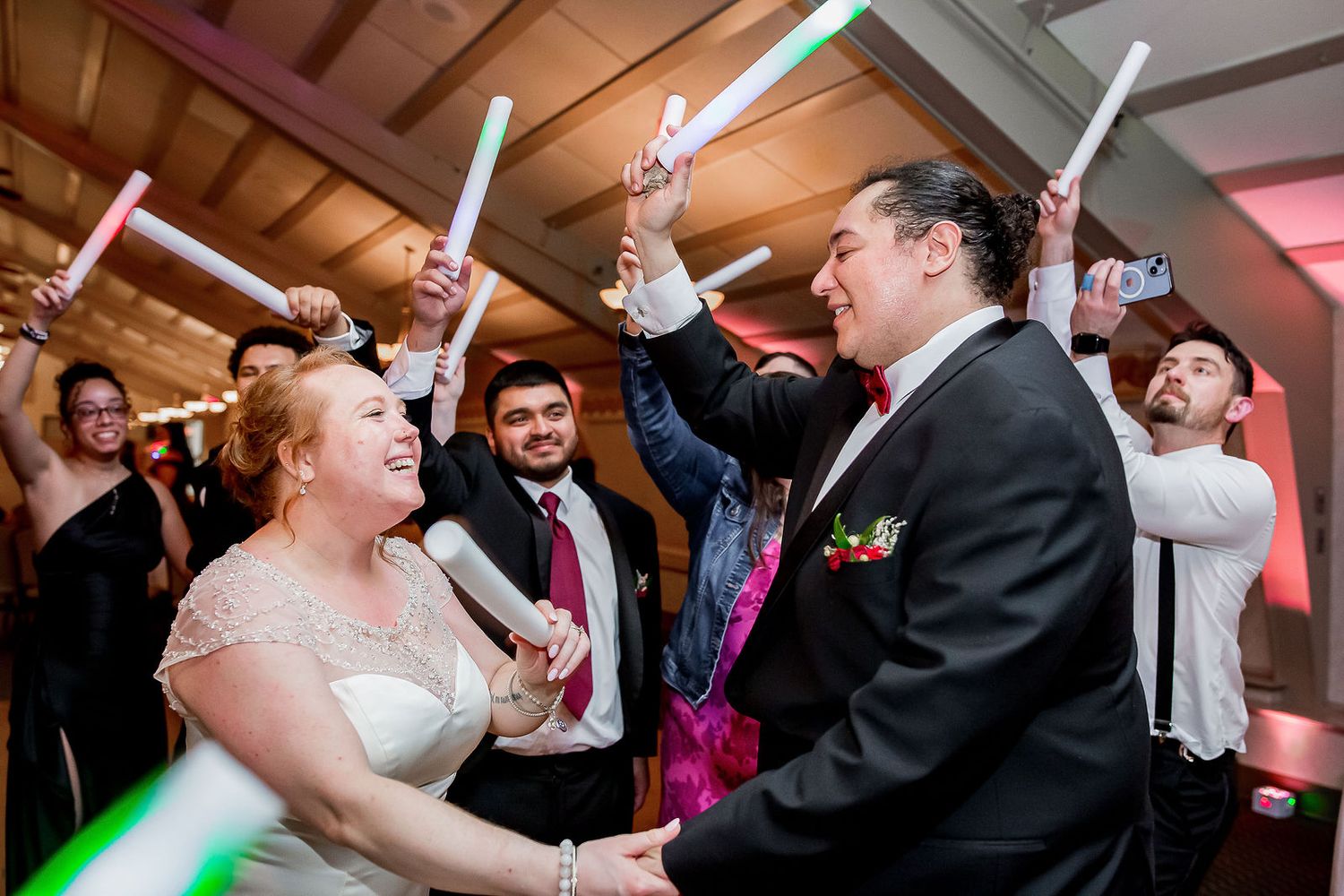 Bride and groom dance with guests holding glowing light sabers at festive wedding reception.