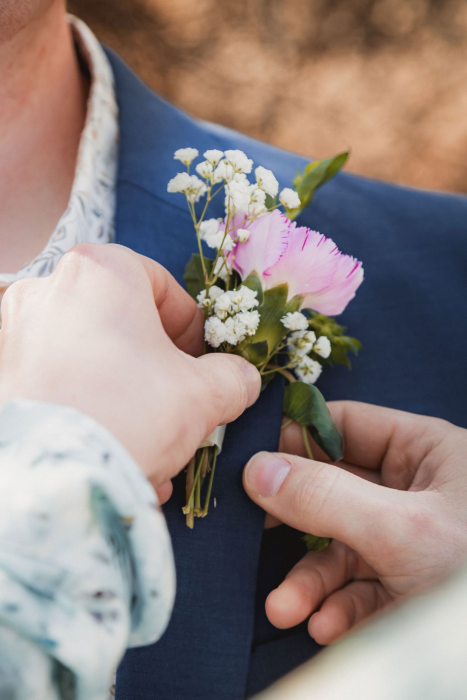 Hands pinning a pink and white floral boutonniere onto a blue suit jacket at a wedding.