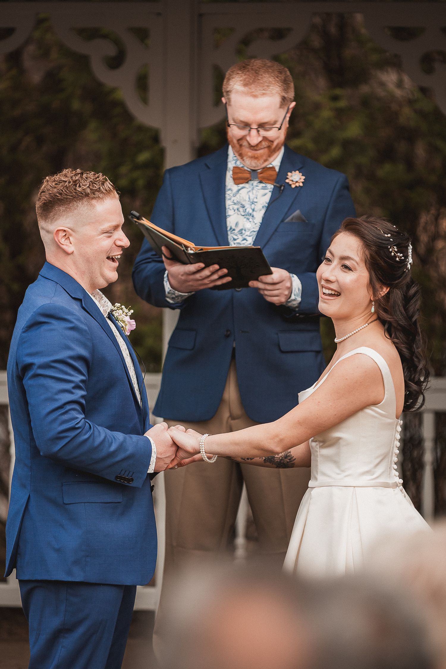 Joyful bride and groom holding hands during outdoor wedding ceremony while officiant in blue suit reads from book.